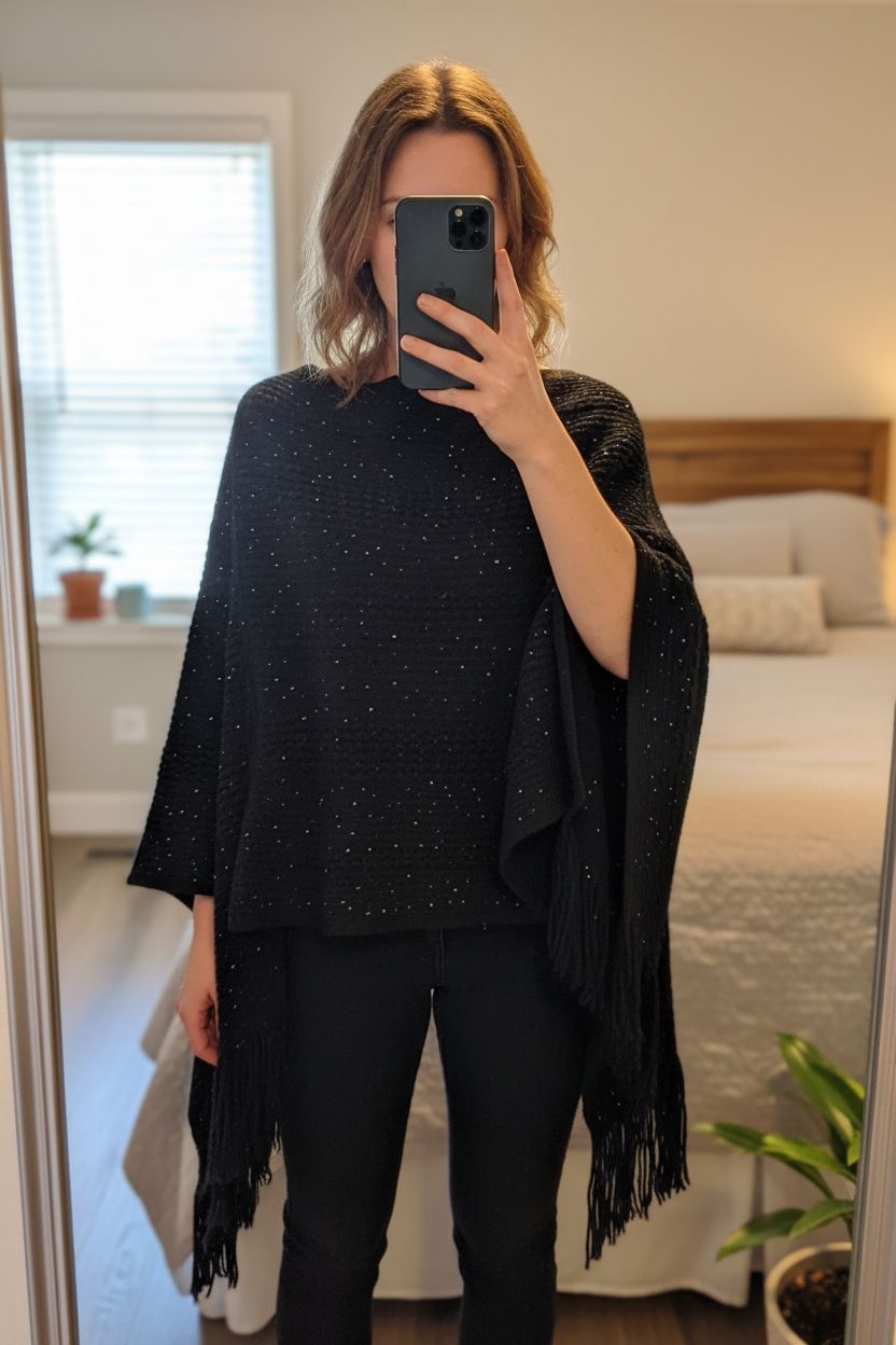 Vintage style black knitted poncho cape free size fringed sequin sparkle witchcore eveningwear standing straight facing the camera, in a softly lit bedroom setting, with gentle natural light filtering through a window.