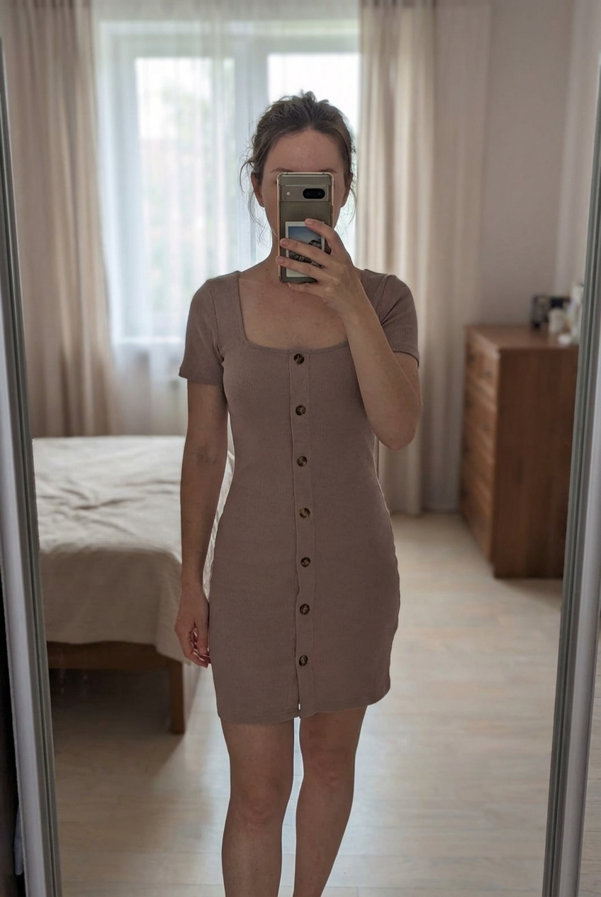 Robe beige avec bouton et col carré debout, face à la caméra, dans une chambre à la lumière douce, avec une lumière naturelle filtrant par la fenêtre.