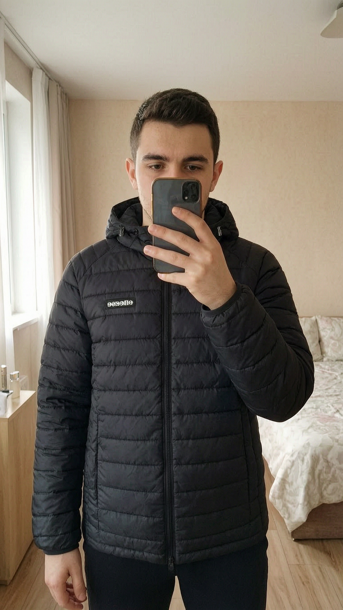 Blouson noir debout, face à la caméra, dans une chambre à la lumière douce, avec une lumière naturelle filtrant par la fenêtre.