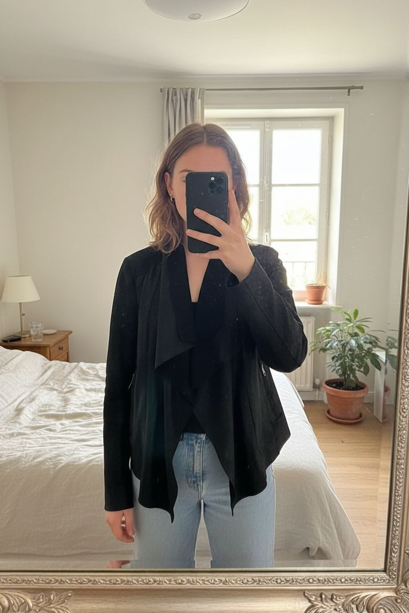 Veste Maje 3 debout, face à la caméra, dans une chambre à la lumière douce, avec une lumière naturelle filtrant par la fenêtre.