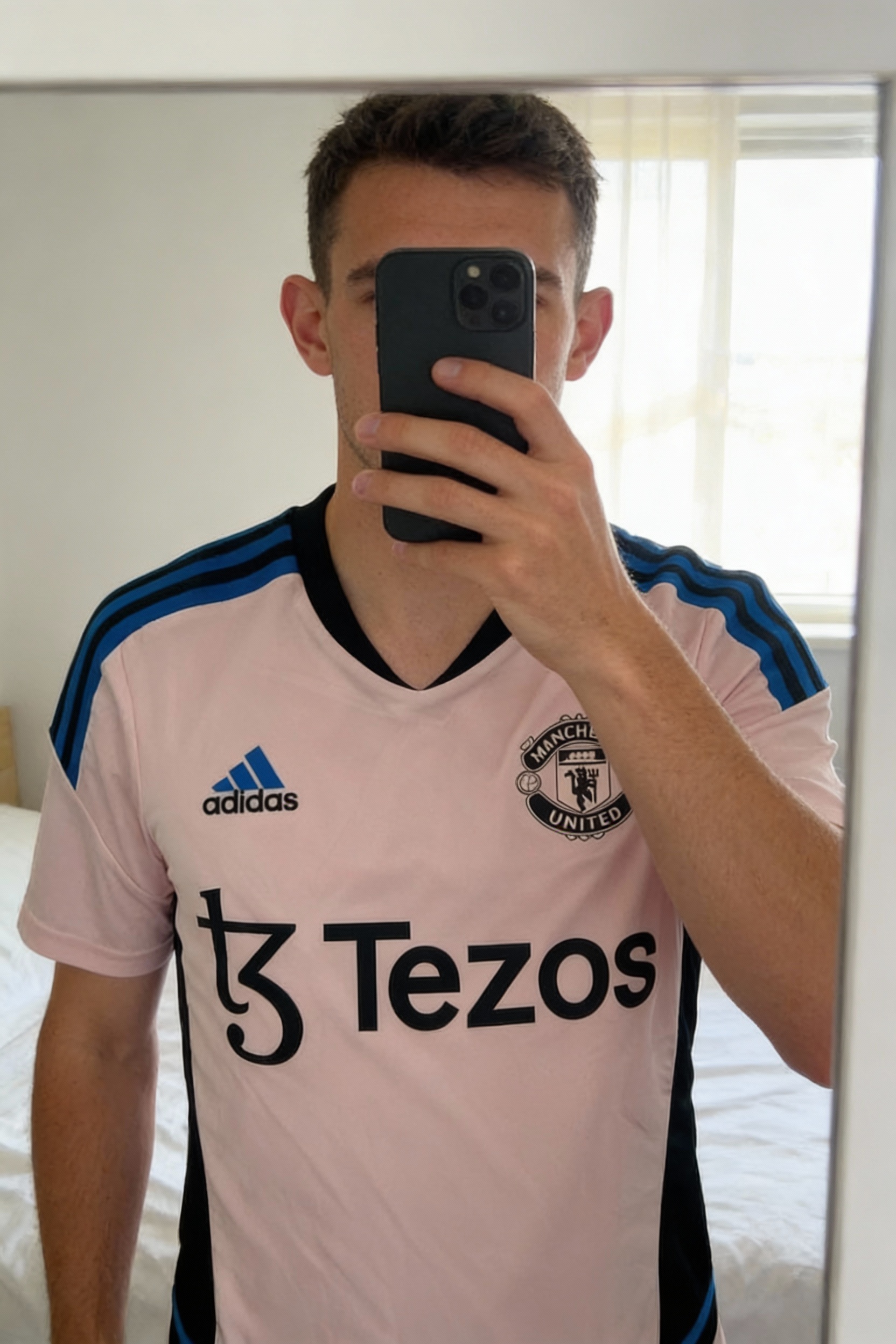 Maillot de football Adidas Manchester United debout, face à la caméra, dans une chambre à la lumière douce, avec une lumière naturelle filtrant par la fenêtre.