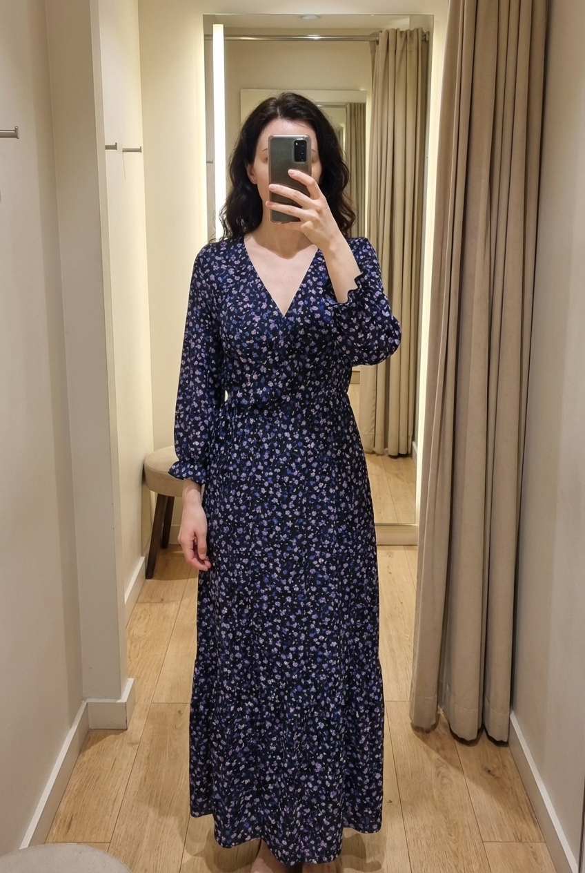 Une personne en Robe longue debout, face à la caméra, dans une cabine d’essayage de style boutique avec un miroir et une décoration neutre.