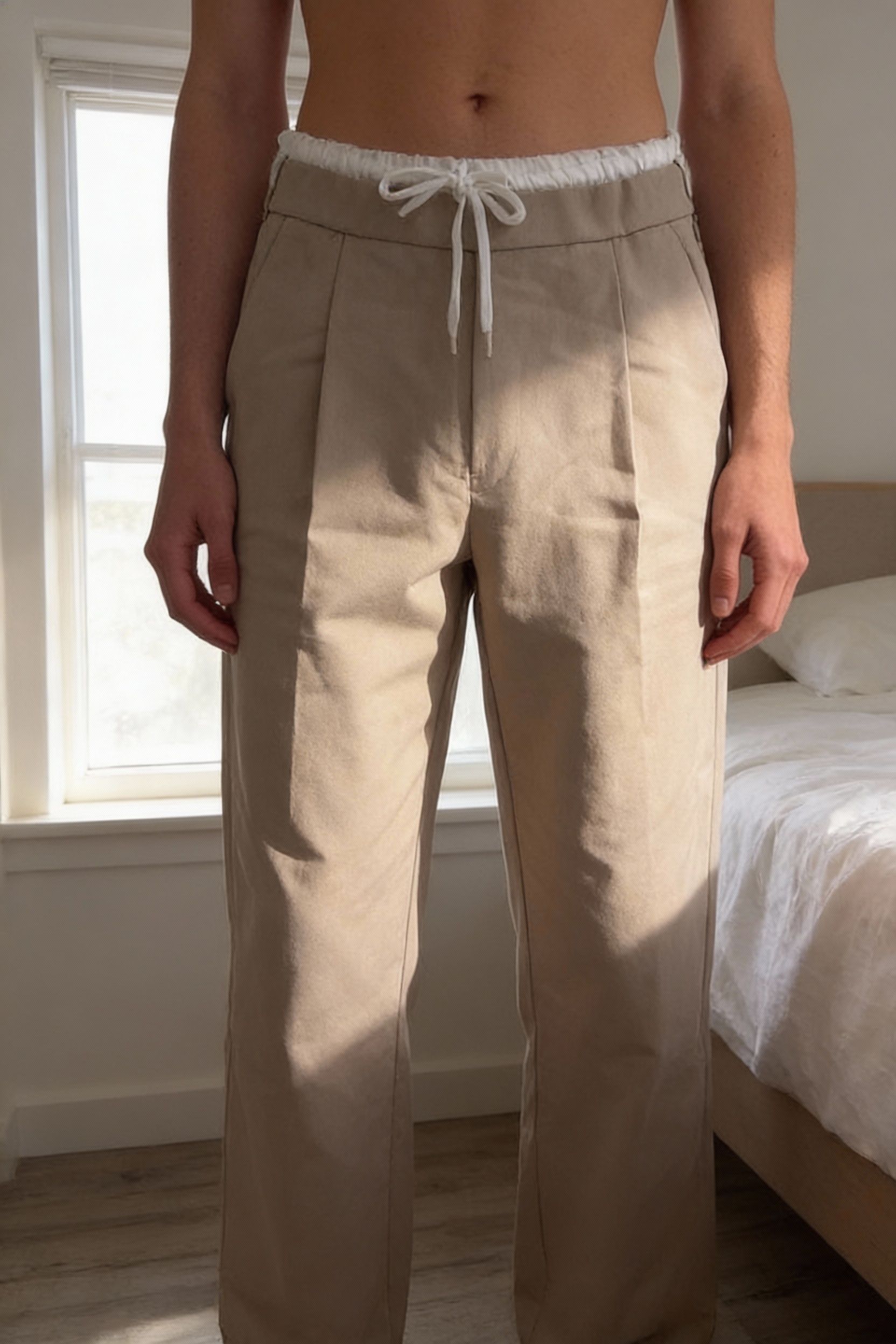 Pantalon beige Zara debout, face à la caméra, dans une chambre à la lumière douce, avec une lumière naturelle filtrant par la fenêtre.