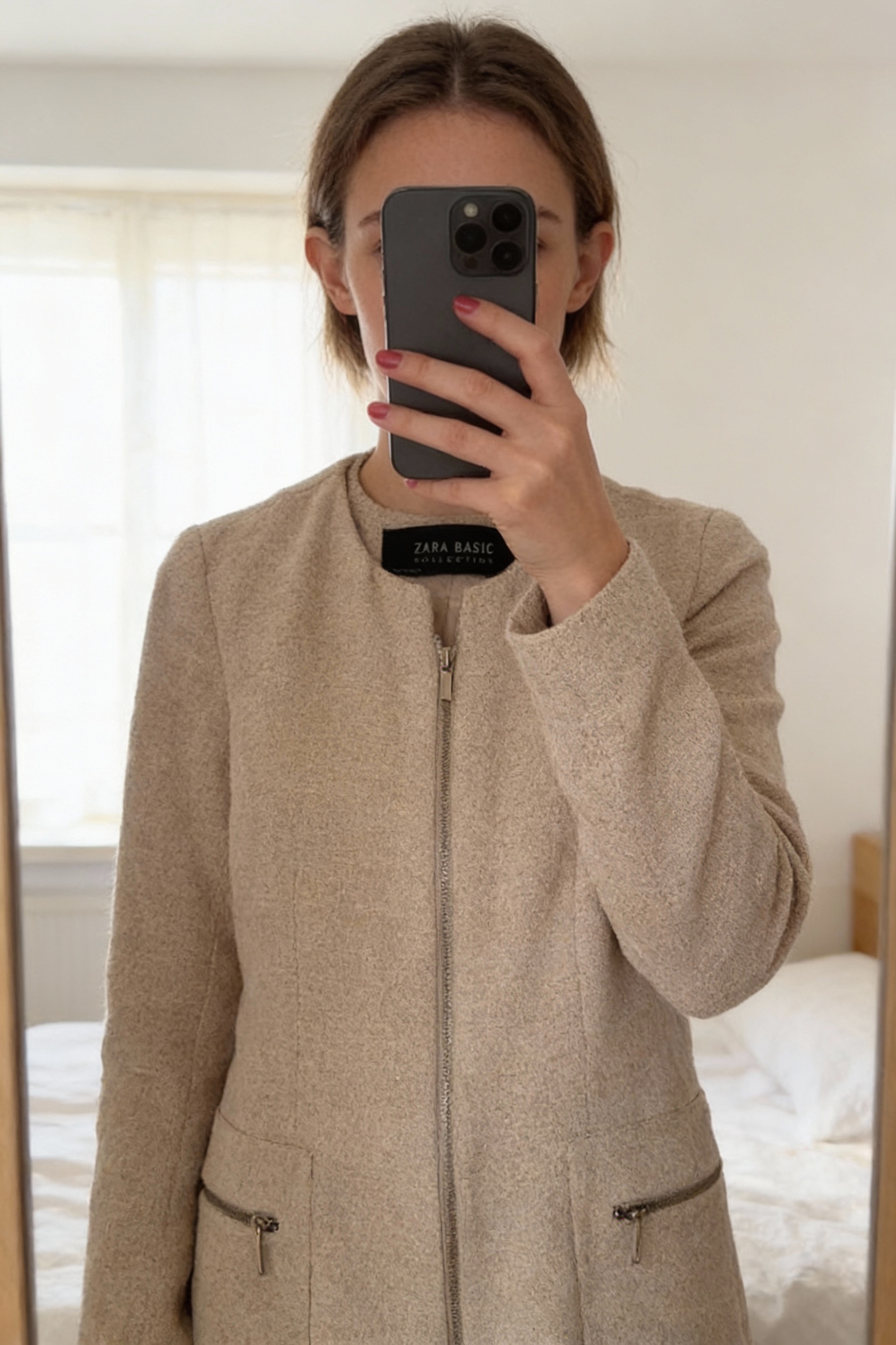 Manteau Zara beige debout, face à la caméra, dans une chambre à la lumière douce, avec une lumière naturelle filtrant par la fenêtre.