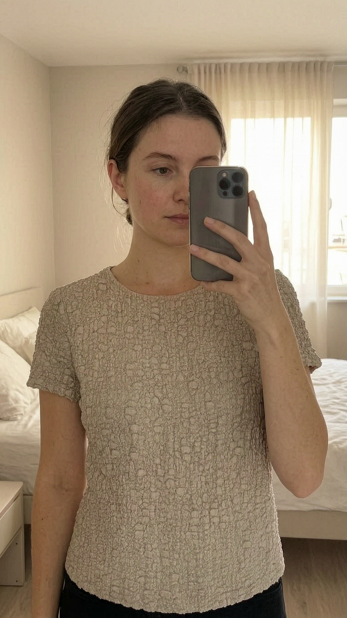 Joli t-shirt beige mango M - Maglietta beige mango Donna M in piedi, rivolto verso la fotocamera, in una camera da letto con luce soffusa e luce naturale che filtra dalla finestra.
