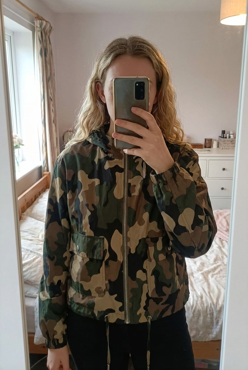 Chaqueta impermeable corta camuflaje de pie, mirando a la cámara, en un dormitorio con luz suave y natural filtrándose por la ventana.