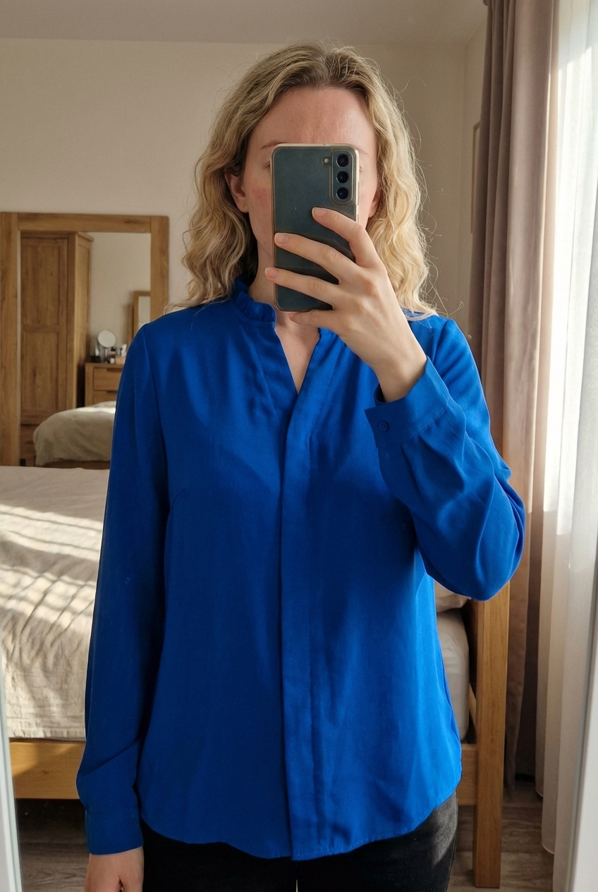 Chemisier bleu debout, face à la caméra, dans une chambre à la lumière douce, avec une lumière naturelle filtrant par la fenêtre.