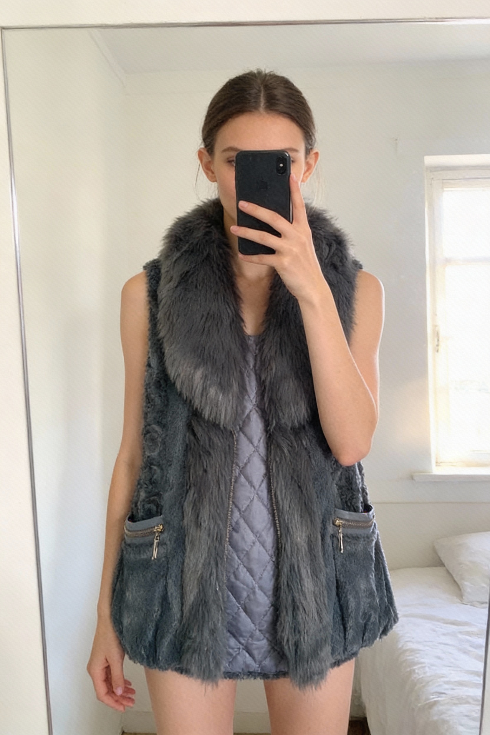 Une personne en Veste sans manches en fausse fourrure debout, face à la caméra, avec une silhouette fine, dans une chambre à la lumière douce, avec une lumière naturelle filtrant par la fenêtre.