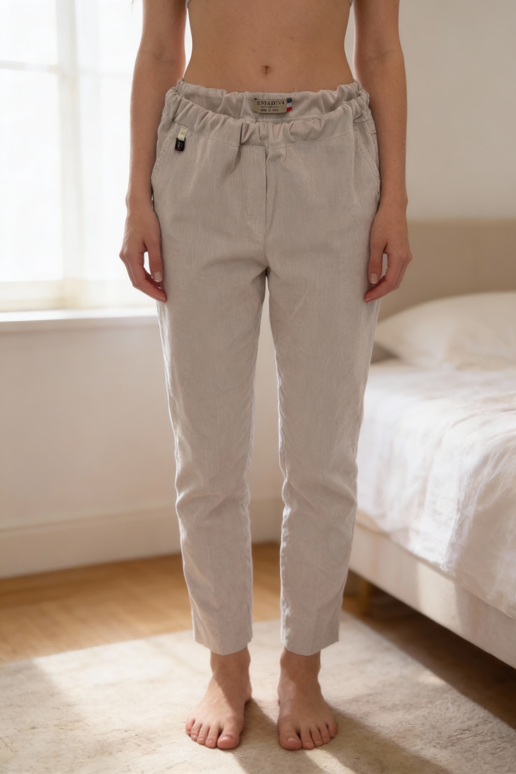 Una persona in Pantalon beige en velours - Pantaloni beige donna M in piedi, rivolto verso la fotocamera, in una camera da letto con luce soffusa e luce naturale che filtra dalla finestra.