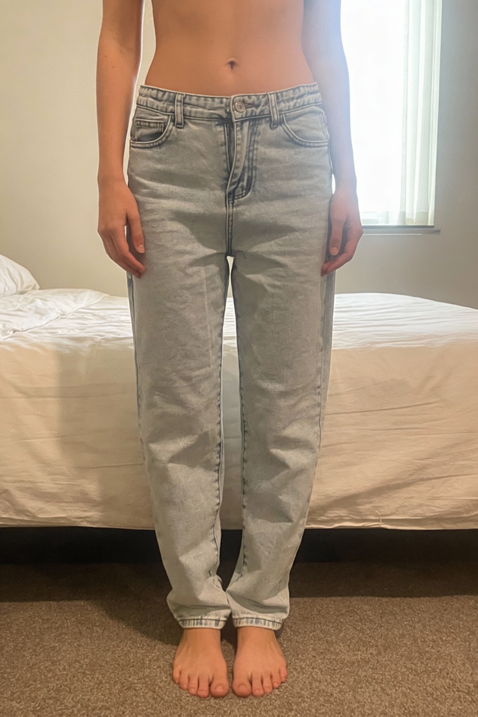 Jean large debout, face à la caméra, dans une chambre à la lumière douce, avec une lumière naturelle filtrant par la fenêtre.