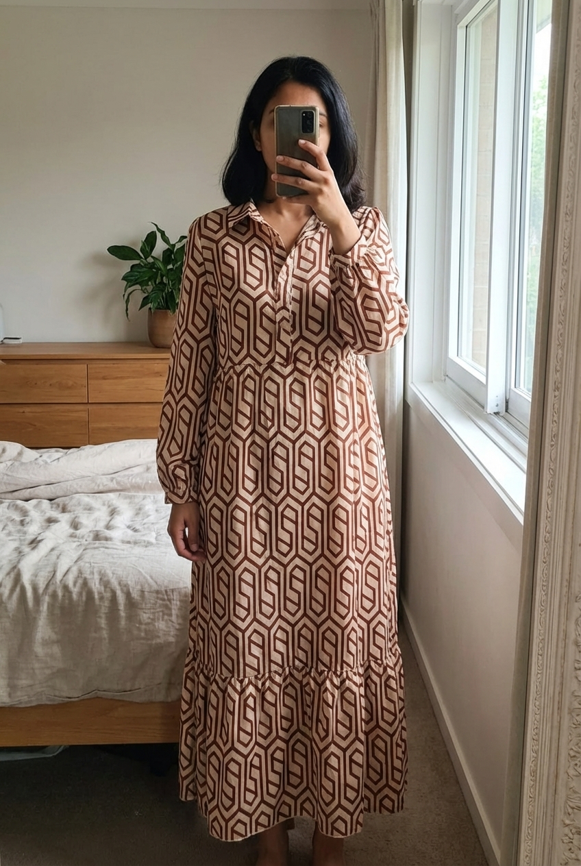 Robe longue pour femme marron debout, face à la caméra, dans une chambre à la lumière douce, avec une lumière naturelle filtrant par la fenêtre.