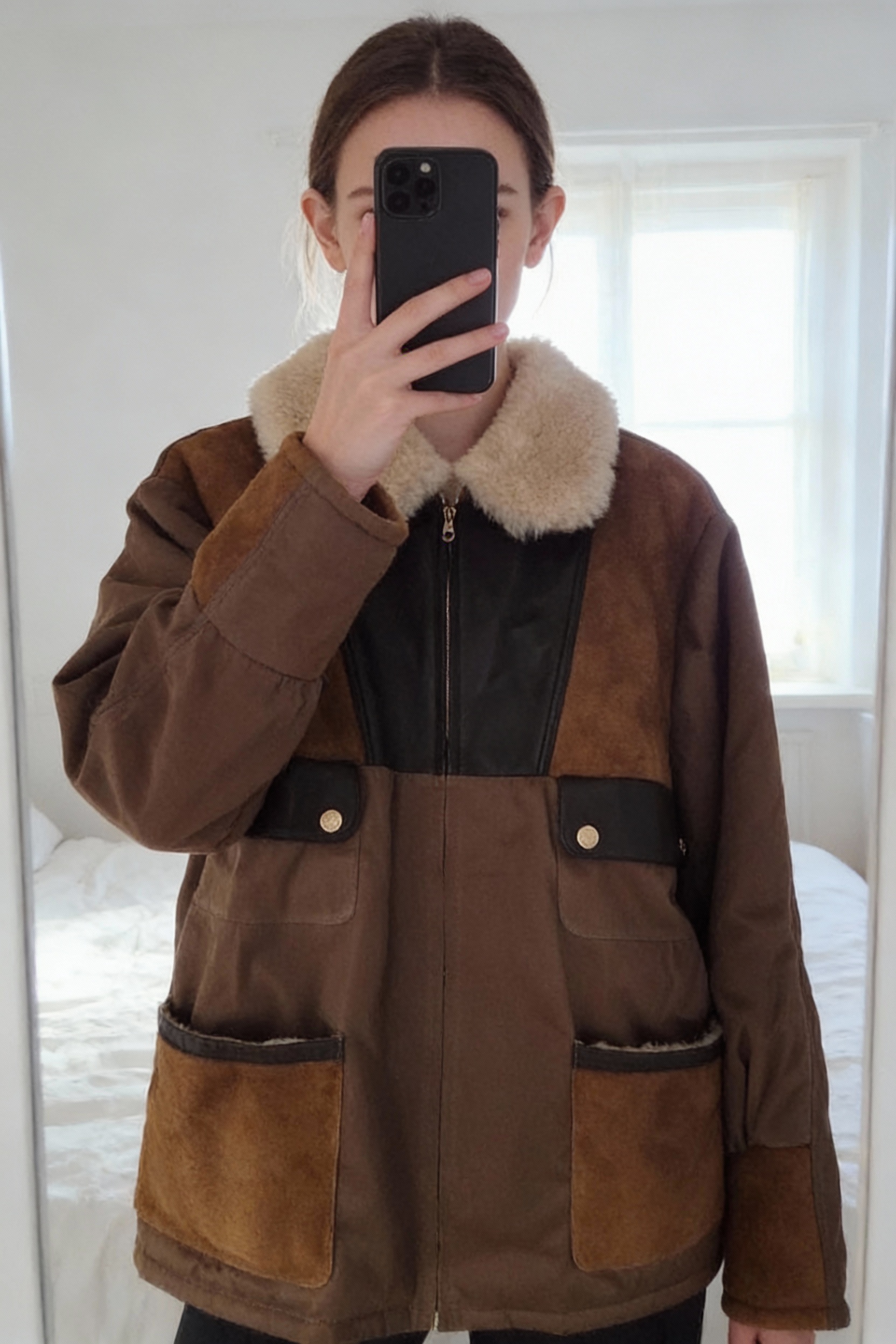 Veste peau lainée Sandro T40 neuf debout, face à la caméra, dans une chambre à la lumière douce, avec une lumière naturelle filtrant par la fenêtre.