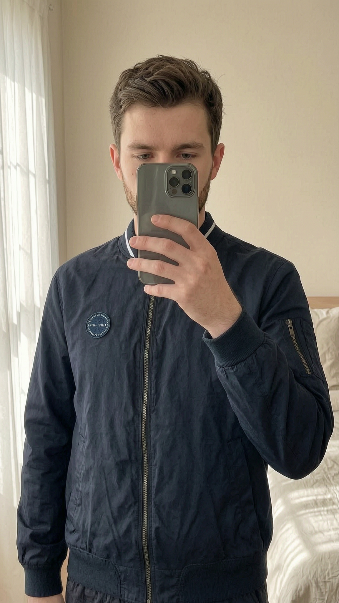 Jack Wills Bomber Jacket Men’s Navy Size M standing straight facing the camera, in a softly lit bedroom setting, with gentle natural light filtering through a window.