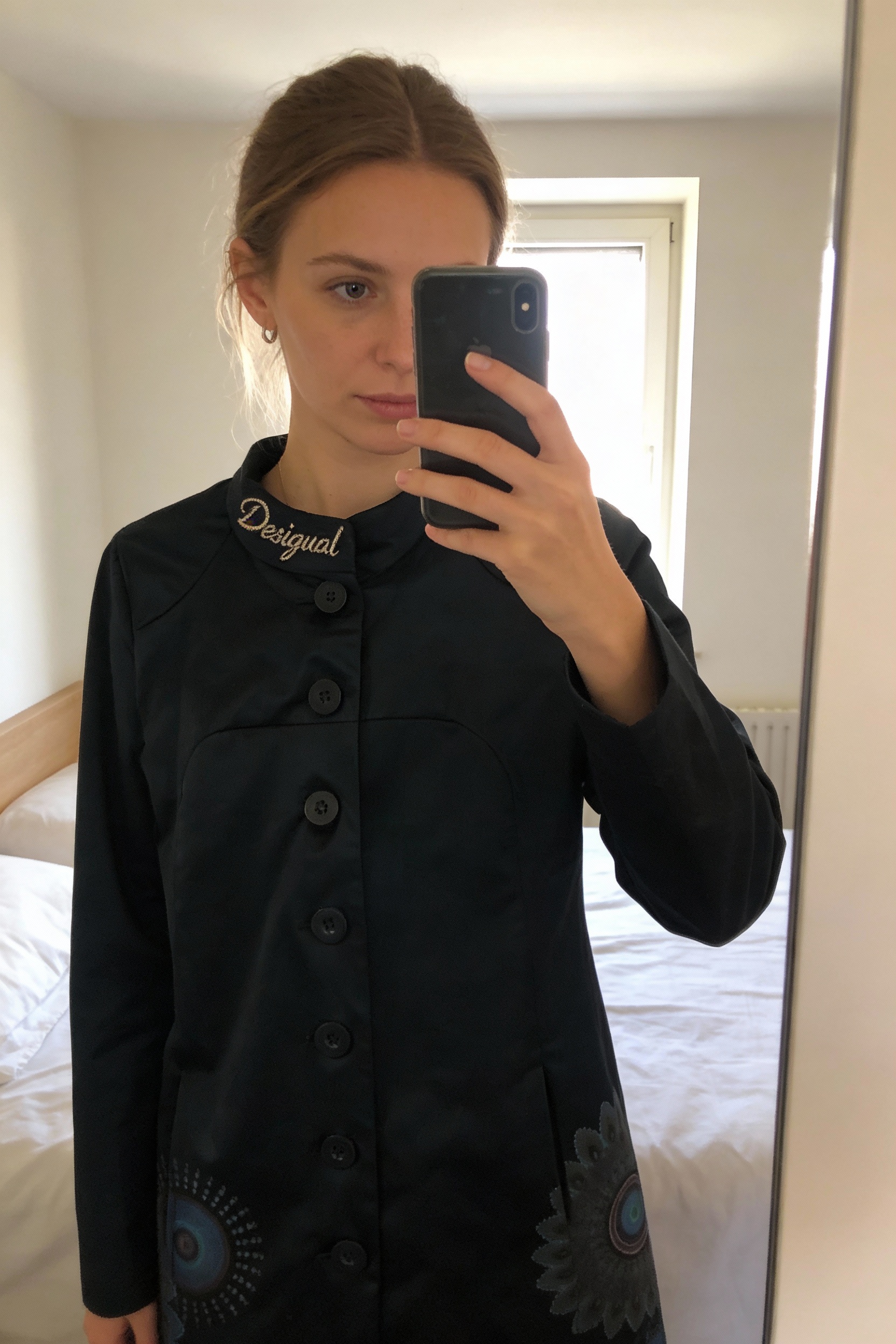 Trench Desigual comme neuf debout, face à la caméra, dans une chambre à la lumière douce, avec une lumière naturelle filtrant par la fenêtre.