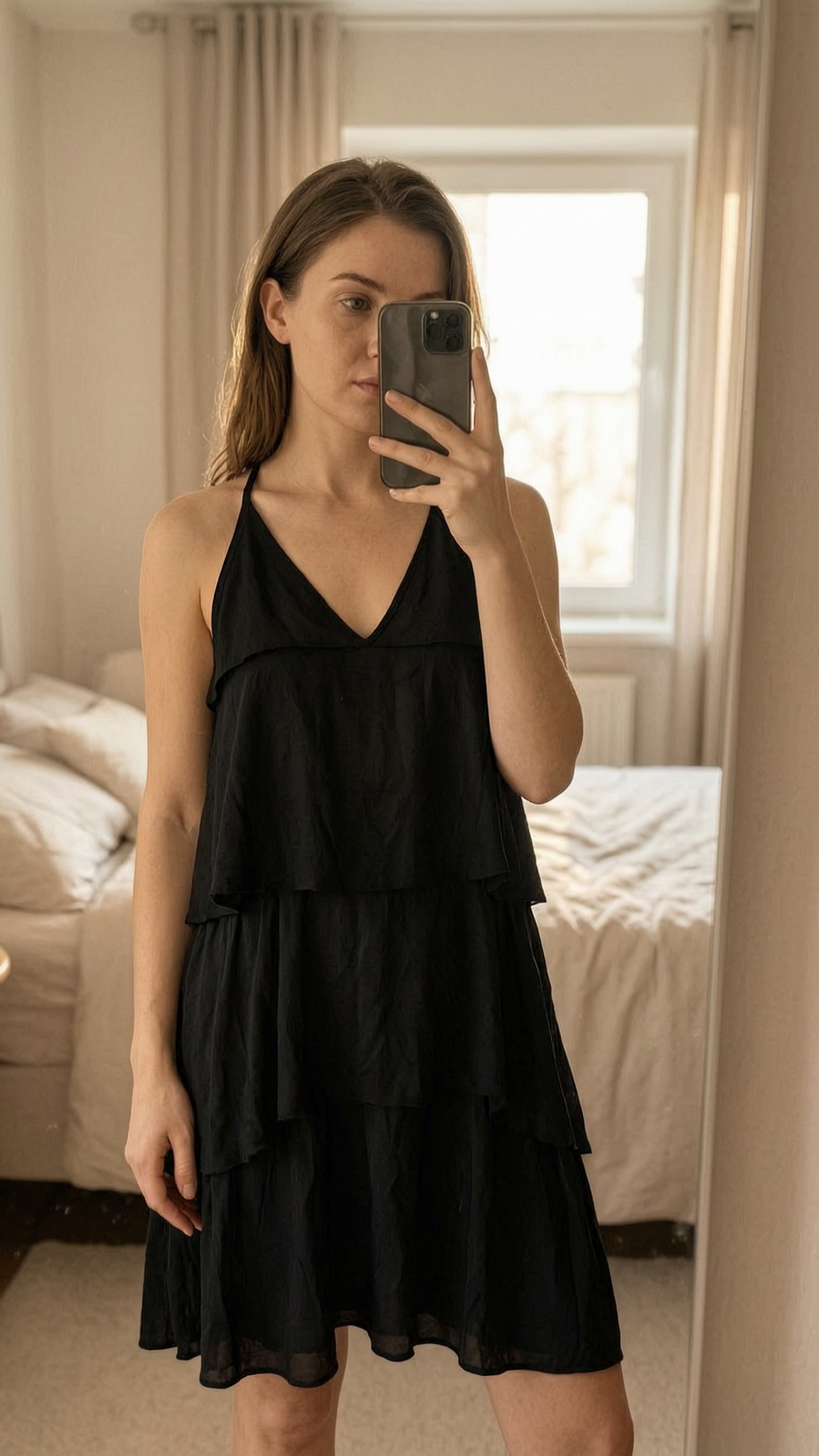 Robe à volant à nouer au cou TU debout, face à la caméra, dans une chambre à la lumière douce, avec une lumière naturelle filtrant par la fenêtre.