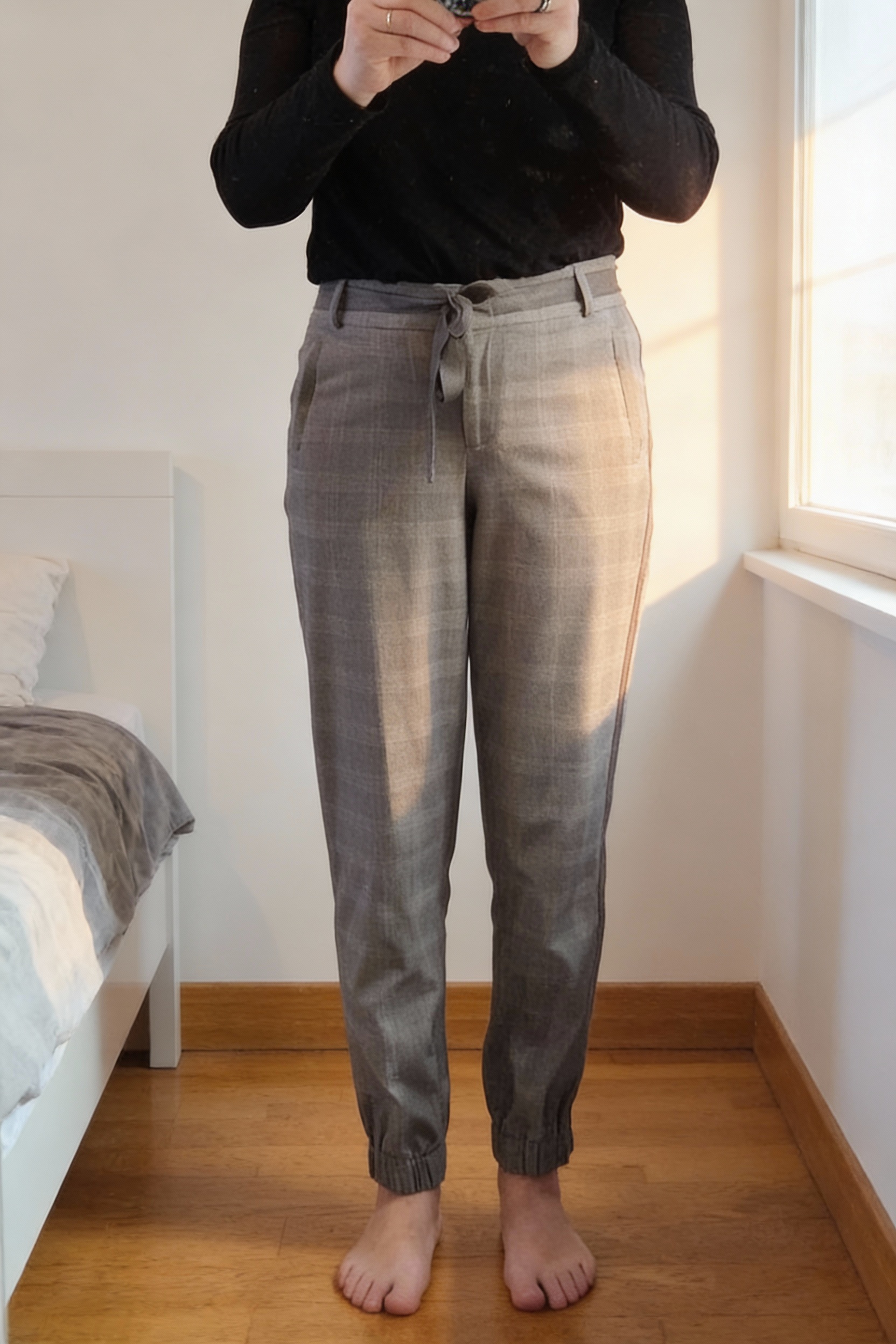 Pantalon gris Prince de Galles debout, face à la caméra, dans une chambre à la lumière douce, avec une lumière naturelle filtrant par la fenêtre.