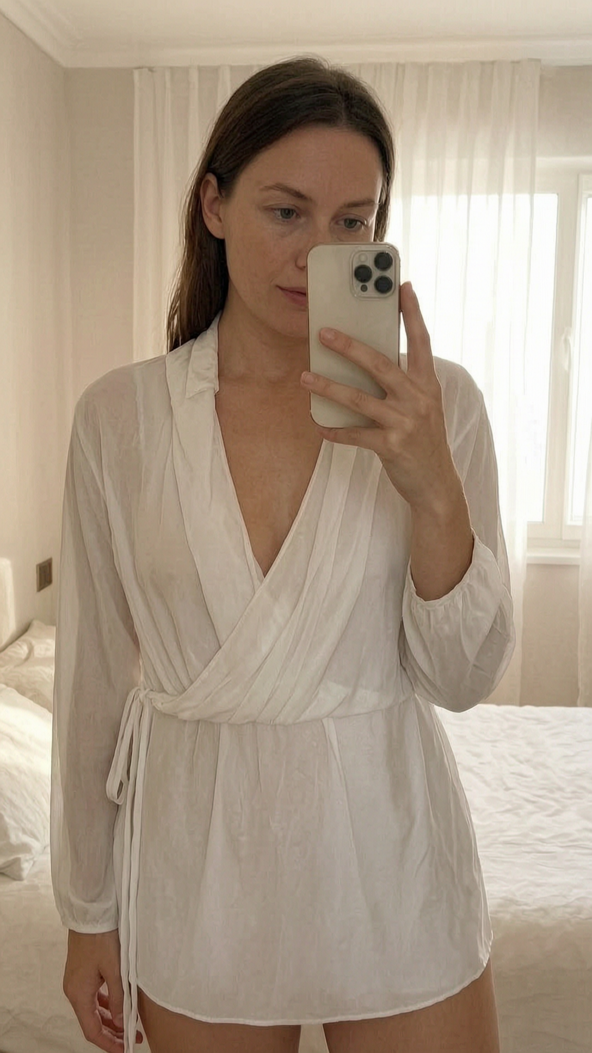 Top cache-cœur blanc – manches longues transparentes – Taille m debout, face à la caméra, dans une chambre à la lumière douce, avec une lumière naturelle filtrant par la fenêtre.