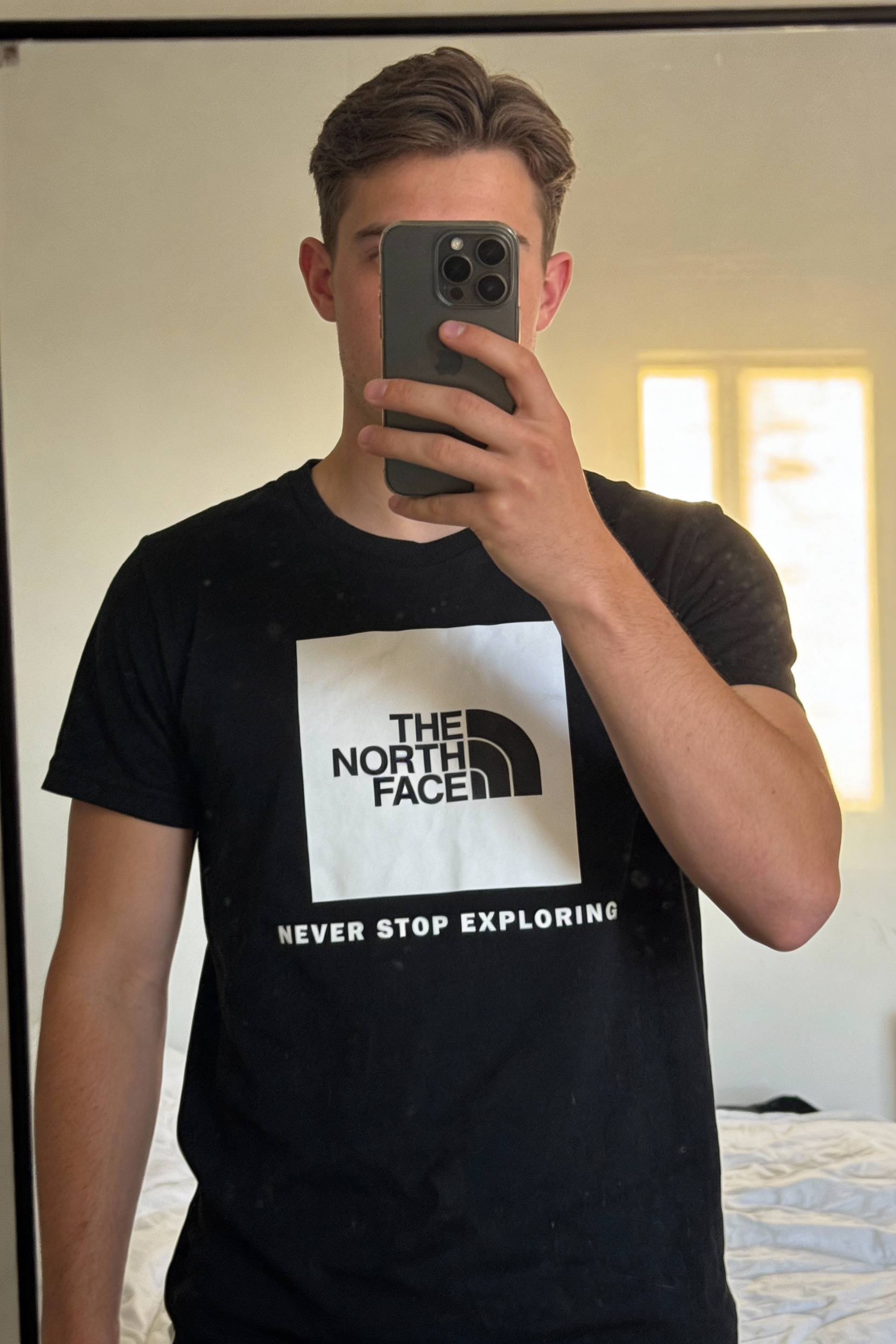 Vends T-Shirt « The North Face » debout, face à la caméra, dans une chambre à la lumière douce, avec une lumière naturelle filtrant par la fenêtre.