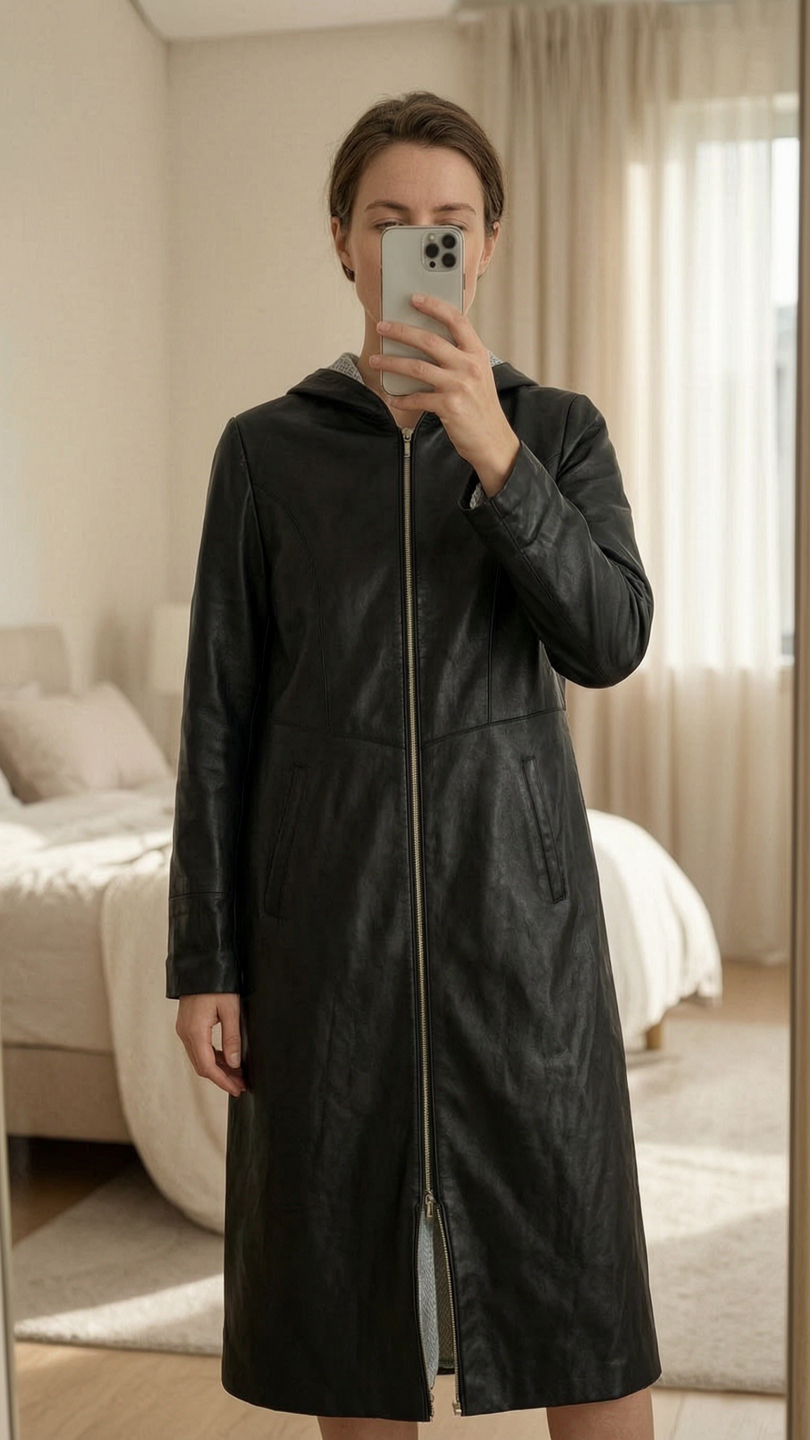 Longue veste zippée à capuche cuir noir debout, face à la caméra, dans une chambre à la lumière douce, avec une lumière naturelle filtrant par la fenêtre.