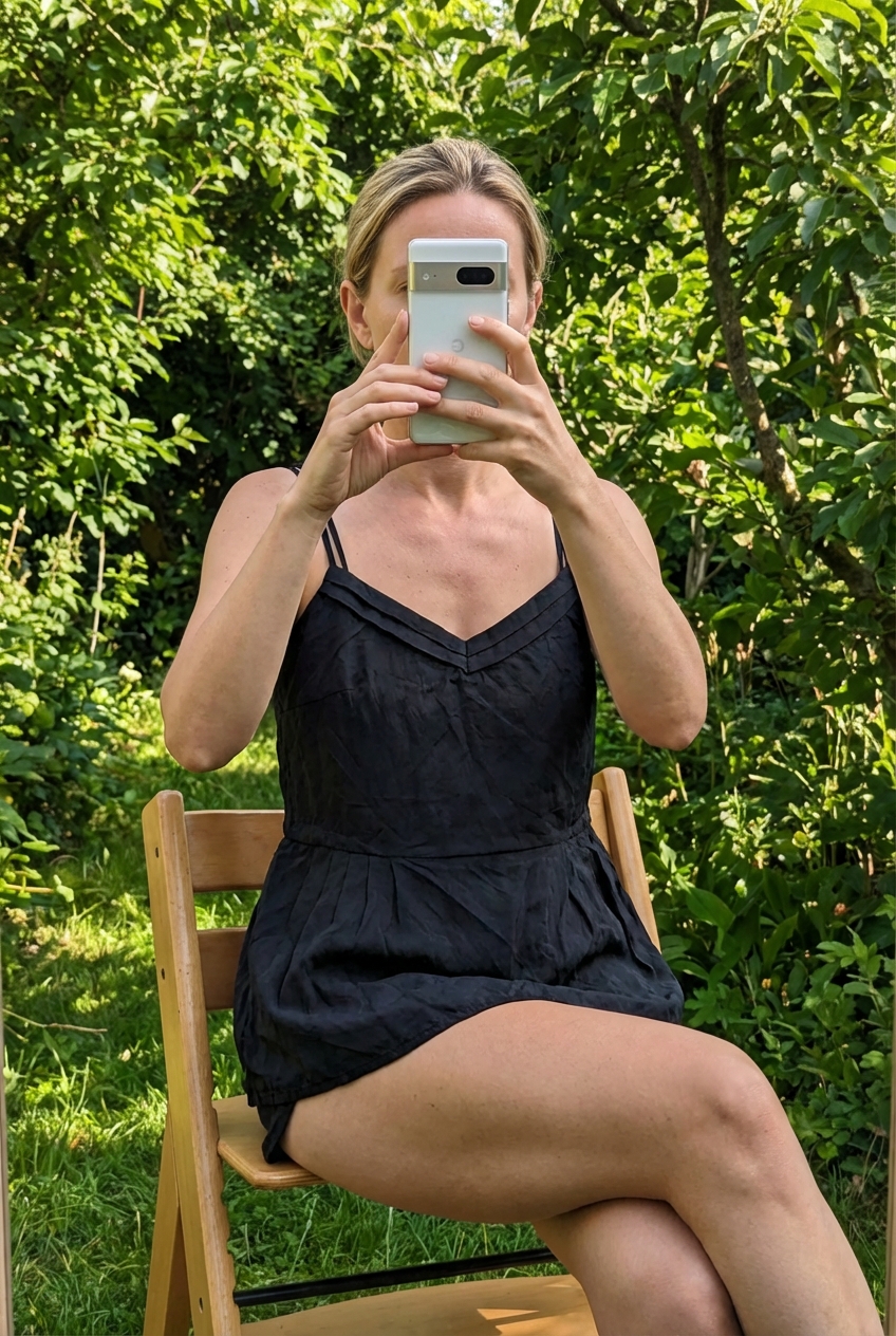 Eine Person in Schwarzes Top von Mint & Berry - schimmert dezent, aber kein Samt! | Gr. 34 auf einem hohen Stuhl sitzend, mit entspannter Haltung, mit schlanker Figur, im Freien, in einer natürlichen Umgebung, umgeben von Grün und sanftem Tageslicht.