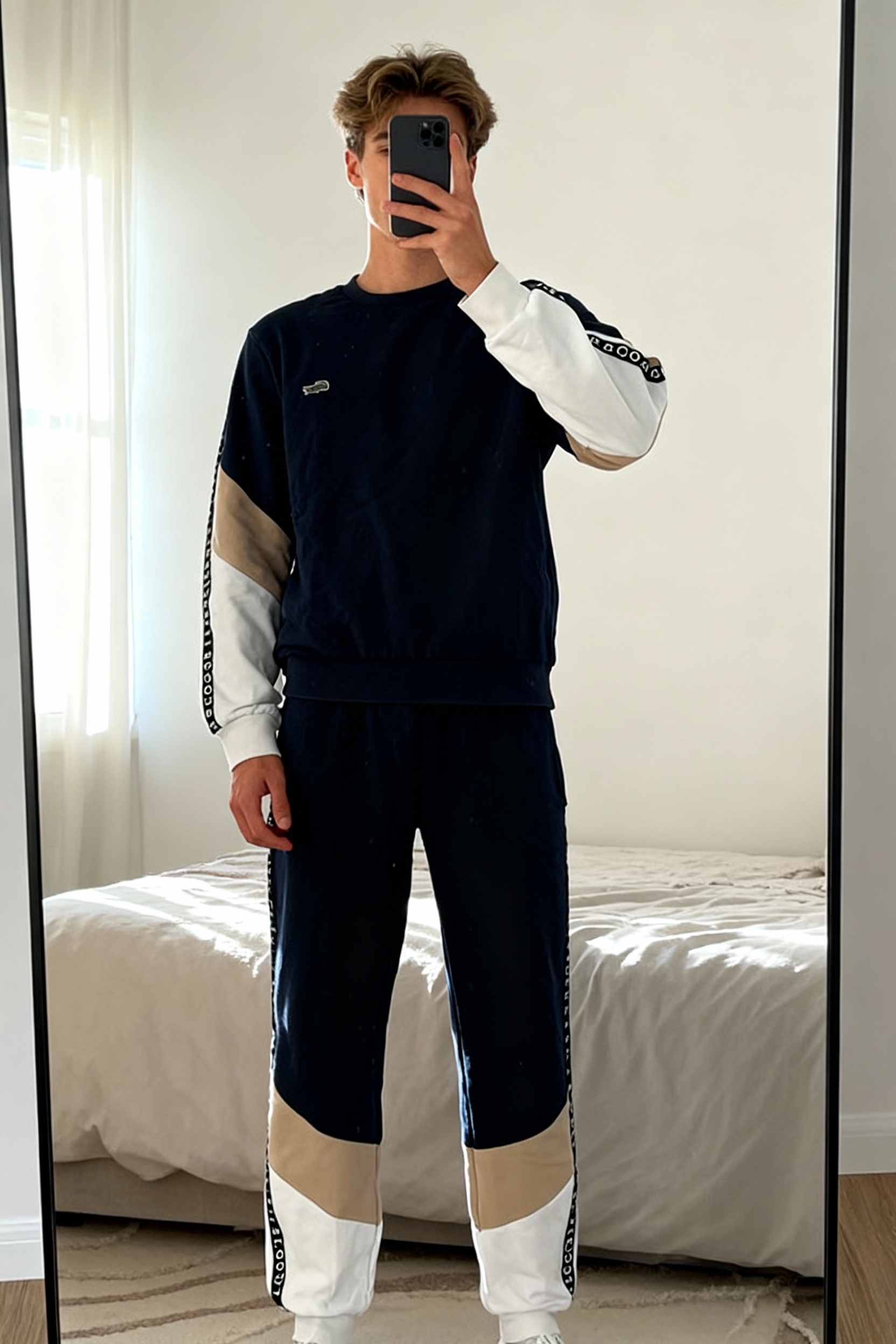 Ensemble de survêtement bande bleu marine, beige, blanc Lacoste debout, face à la caméra, dans une chambre à la lumière douce, avec une lumière naturelle filtrant par la fenêtre.