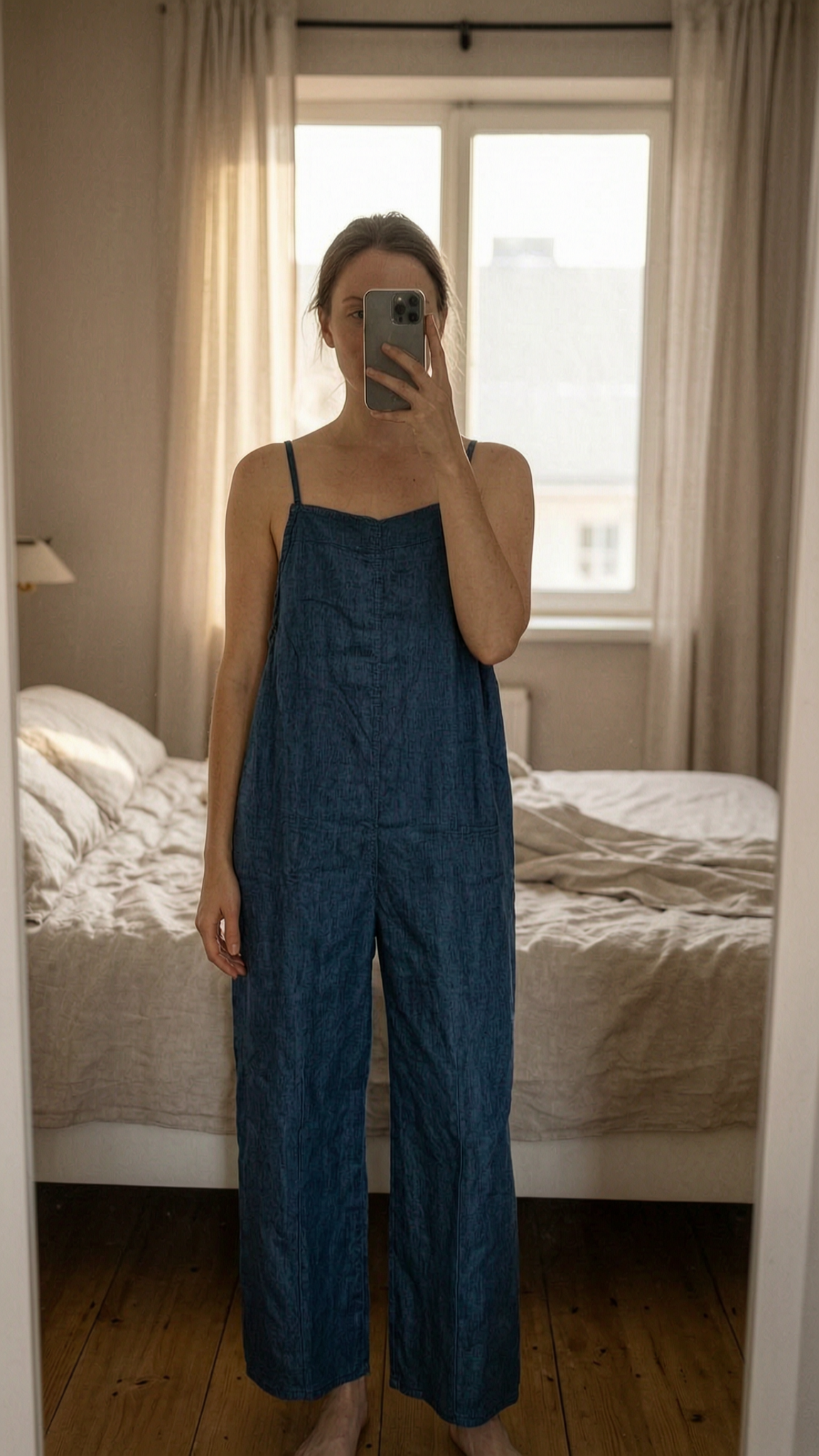 Combi-pantalon debout, face à la caméra, dans une chambre à la lumière douce, avec une lumière naturelle filtrant par la fenêtre.
