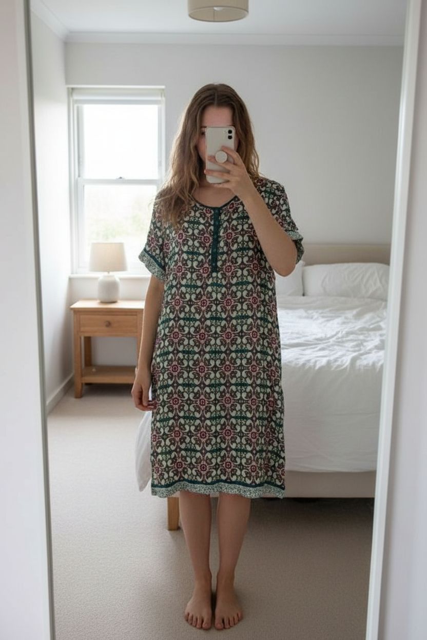 Robe à motifs femme M - Abito estate Donna M in piedi, rivolto verso la fotocamera, in una camera da letto con luce soffusa e luce naturale che filtra dalla finestra.
