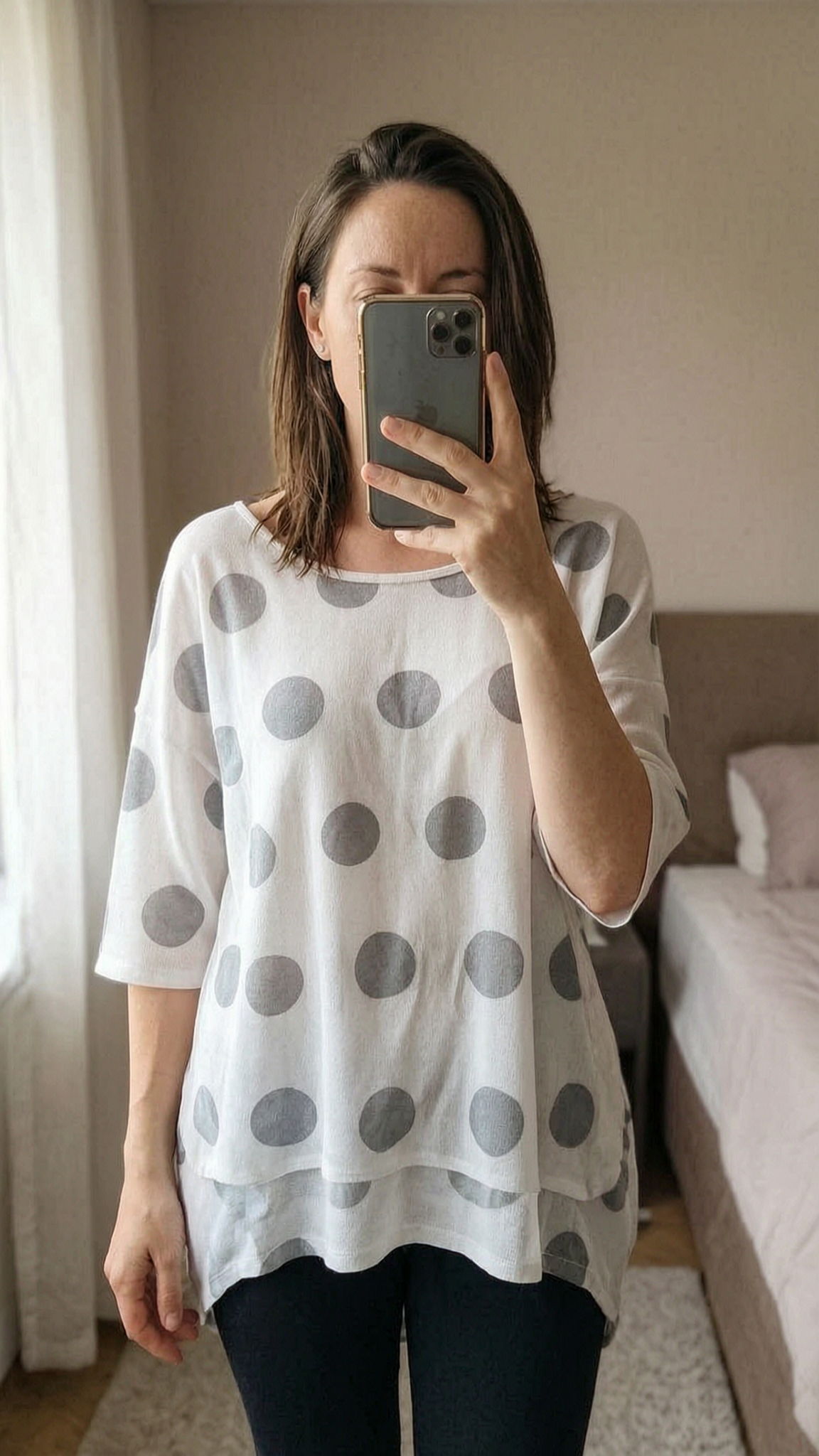 Pull blanc pois debout, face à la caméra, dans une chambre à la lumière douce, avec une lumière naturelle filtrant par la fenêtre.