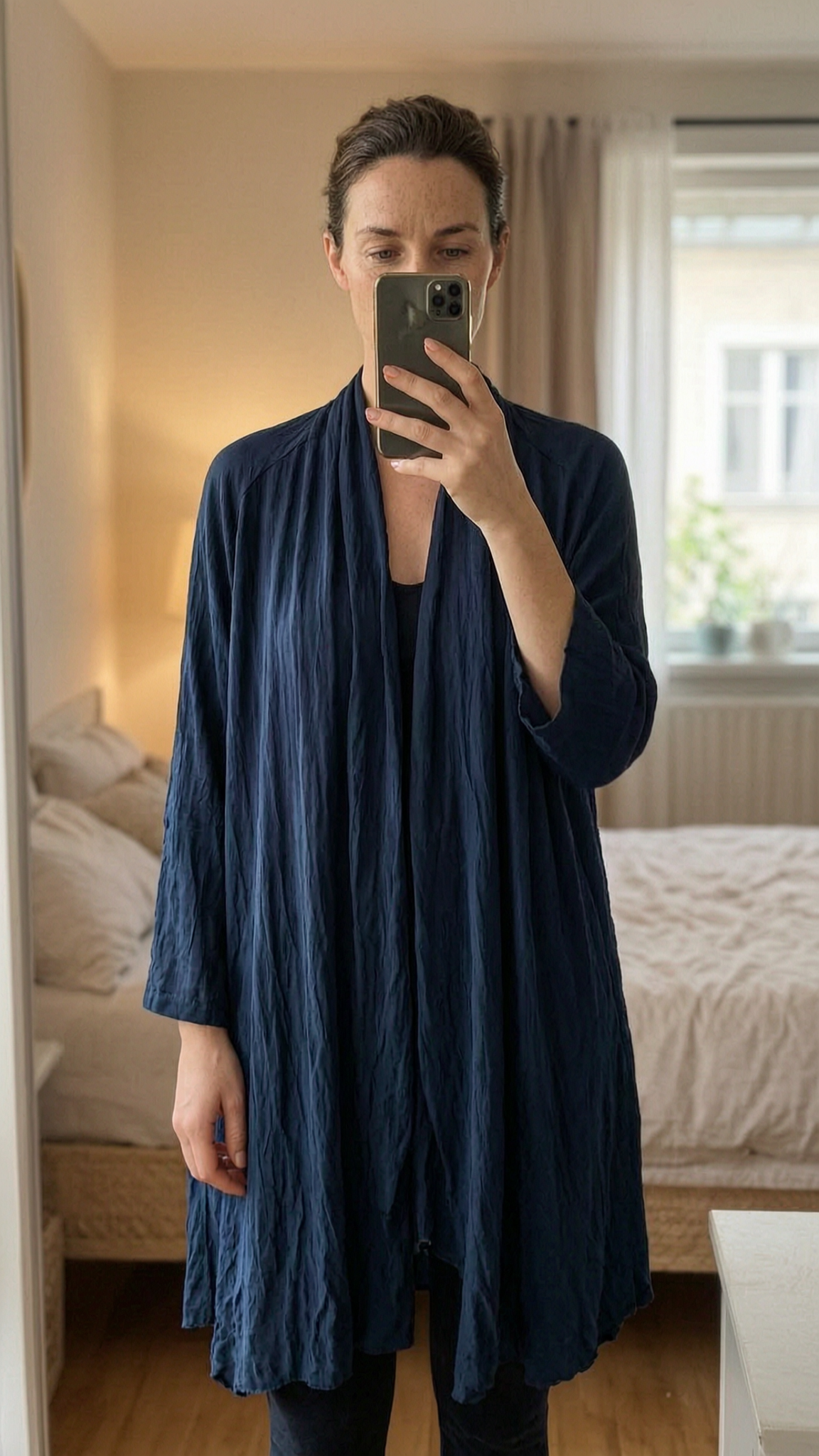 Long gilet fluide léger M - cardigan gilet Donna ragno in piedi, rivolto verso la fotocamera, in una camera da letto con luce soffusa e luce naturale che filtra dalla finestra.