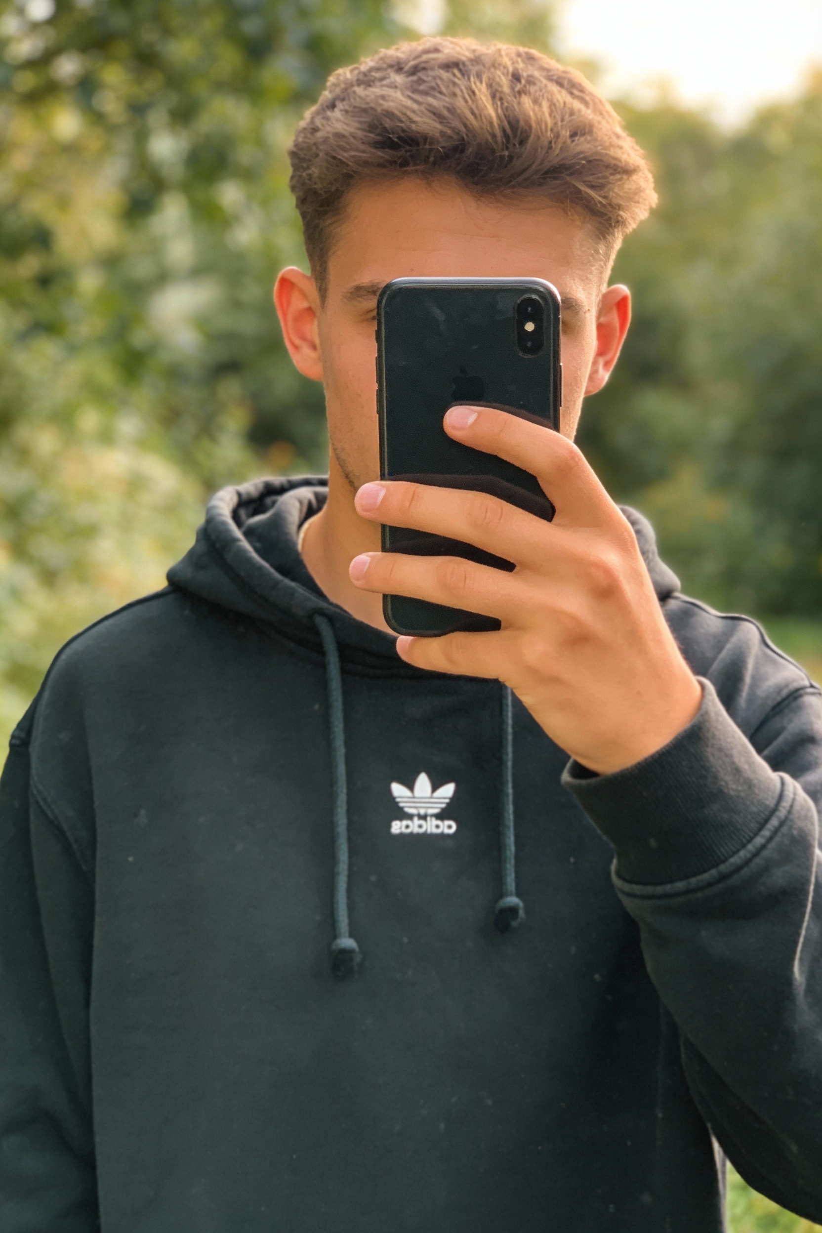 Una persona con Sudadera vintage Adidas con capucha de pie, mirando a la cámara, al aire libre, en un entorno natural rodeado de vegetación y luz suave.