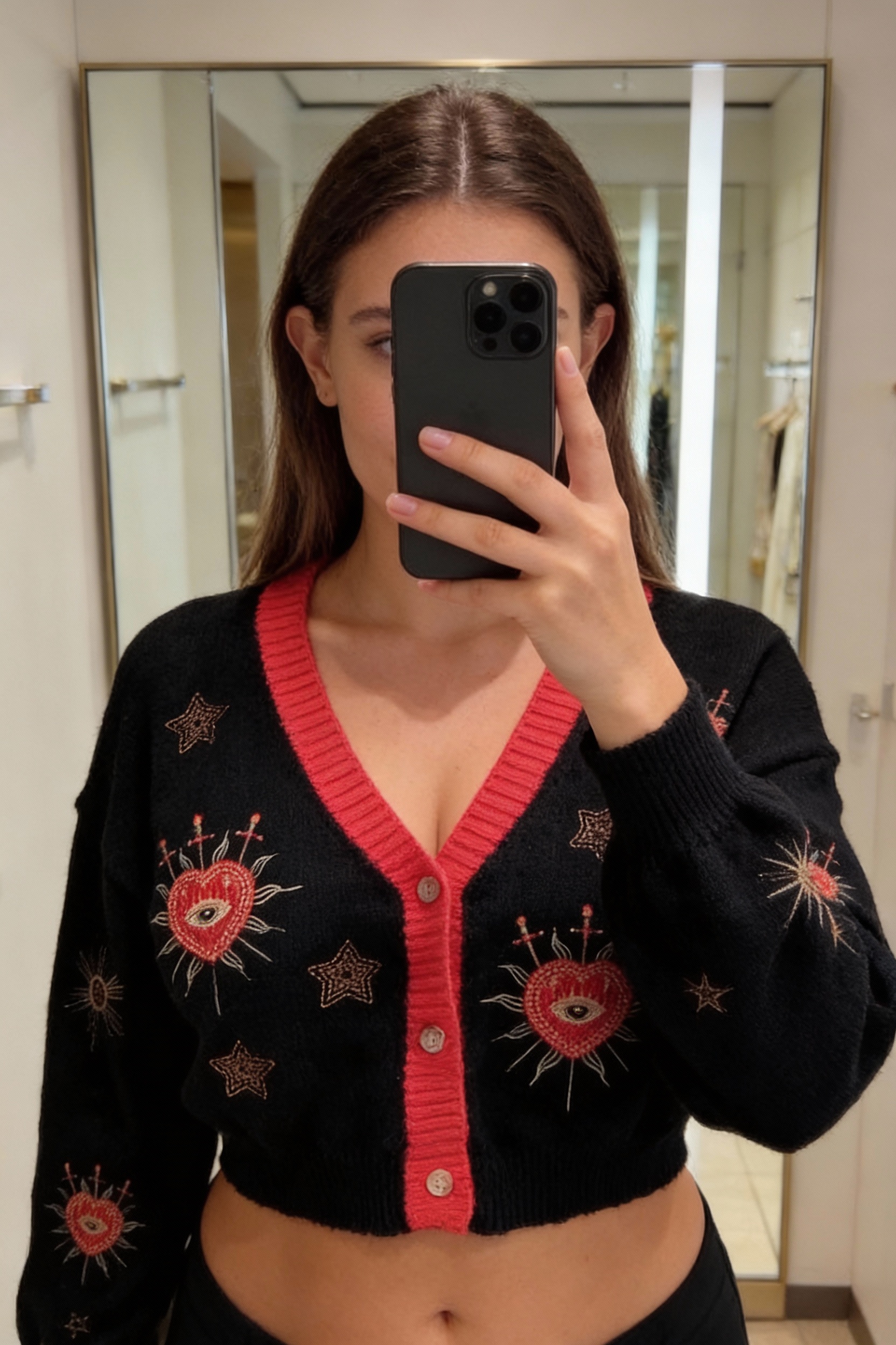 Une personne en Gilet Cardigan tricoté cœurs étoiles debout, face à la caméra, avec une silhouette légèrement plus ronde, dans une cabine d’essayage de style boutique avec un miroir et une décoration neutre.