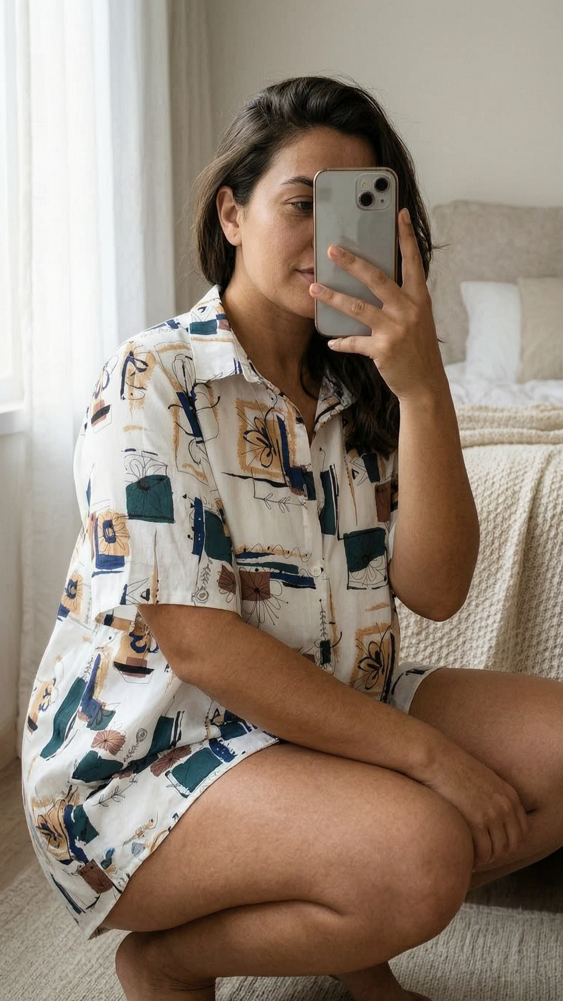 Eine Person in Unisex Hemd mit Printmuster elegant in die Hocke gehend, um den Fall und die Bewegung des Kleidungsstücks zu zeigen, mit etwas kurvigeren Körperformen, in einem sanft beleuchteten Schlafzimmer, mit natürlichem Licht, das durch ein Fenster fällt.