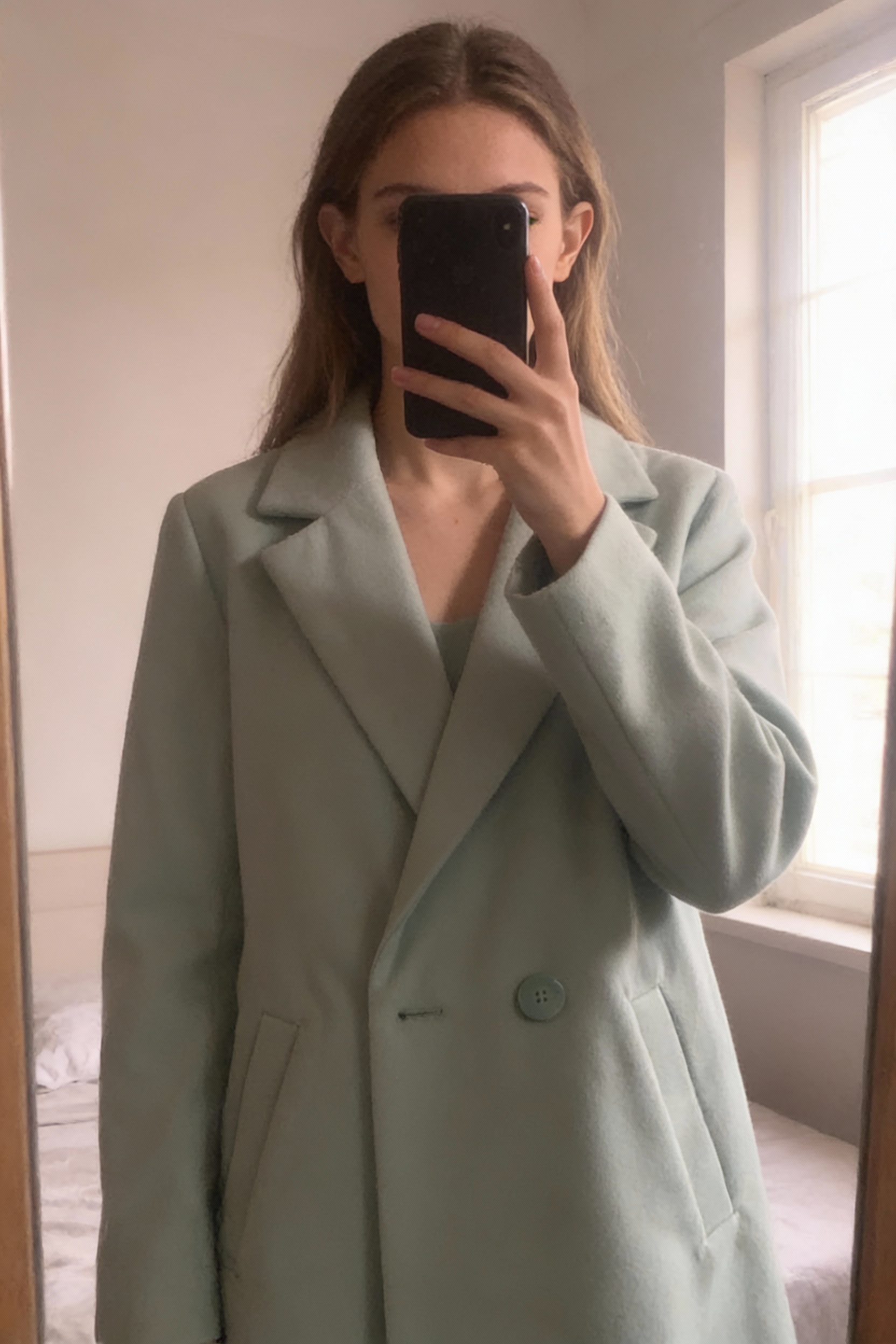 manteau blazer vert pistache cache cache taille XS debout, face à la caméra, dans une chambre à la lumière douce, avec une lumière naturelle filtrant par la fenêtre.