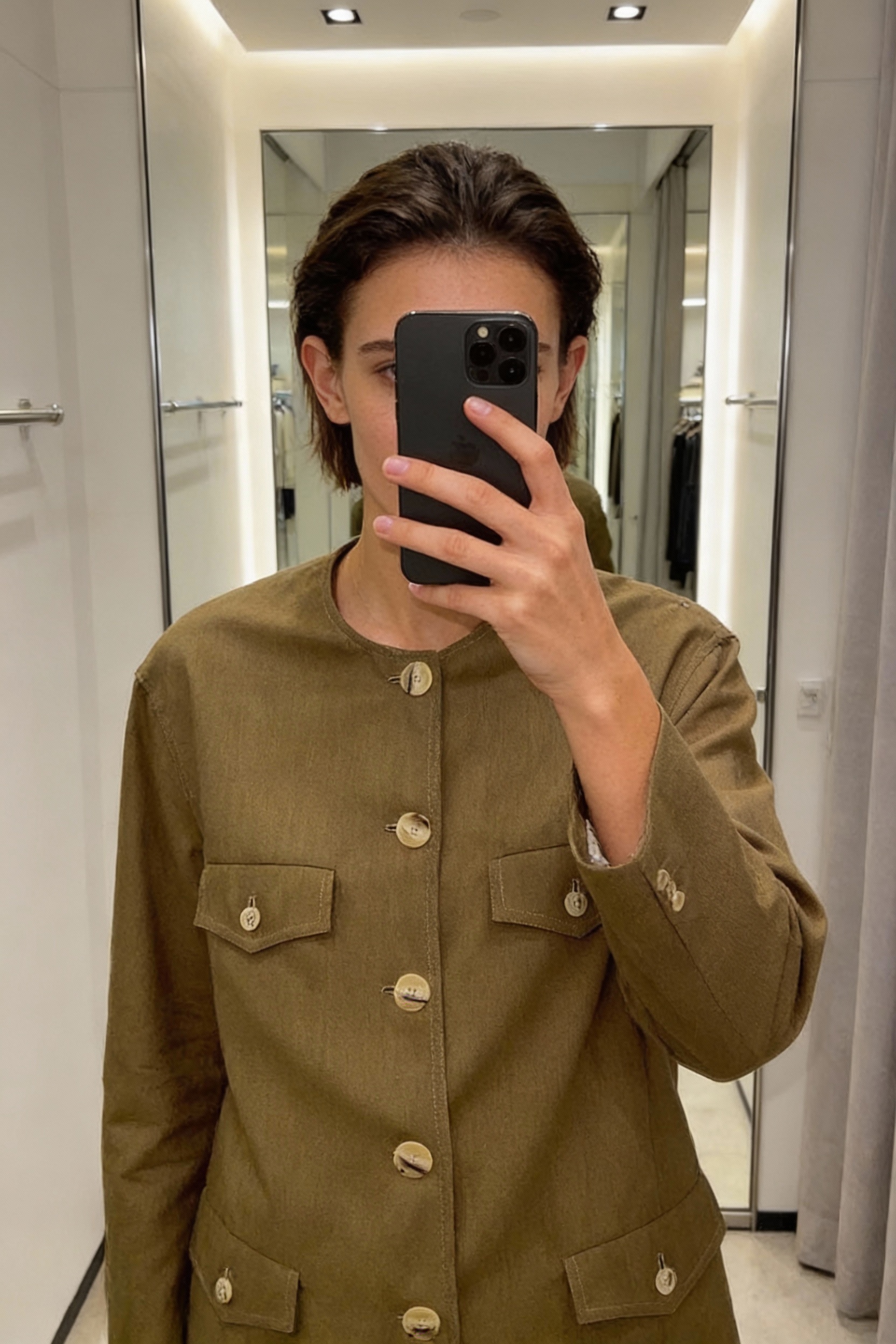 Una persona in Giacca Strutturata Minimal - Lana Vergine - Taglia 44 Cod.3165 in piedi, rivolto verso la fotocamera, in un camerino in stile boutique con specchio e decorazioni neutre.