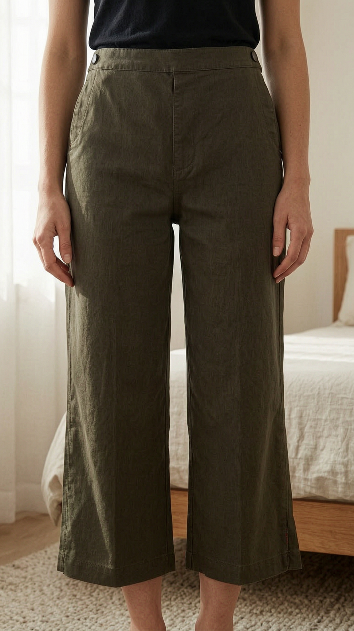 Pantacourt cop.copine taille 40 kaki debout, face à la caméra, dans une chambre à la lumière douce, avec une lumière naturelle filtrant par la fenêtre.