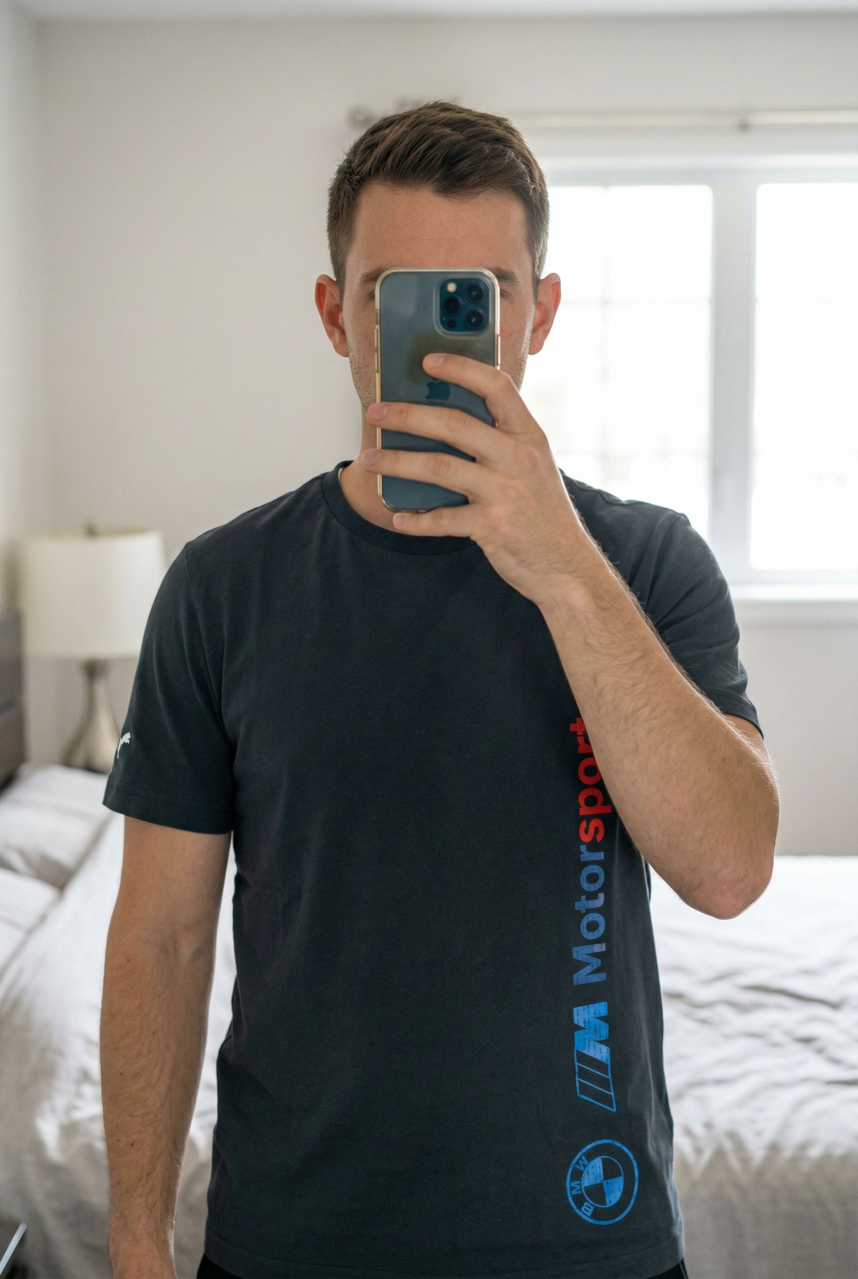 Teeshirt puma bmw taille M debout, face à la caméra, dans une chambre à la lumière douce, avec une lumière naturelle filtrant par la fenêtre.