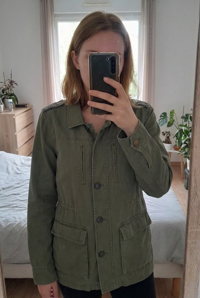 Veste légère kaki S debout, face à la caméra, dans une chambre à la lumière douce, avec une lumière naturelle filtrant par la fenêtre.