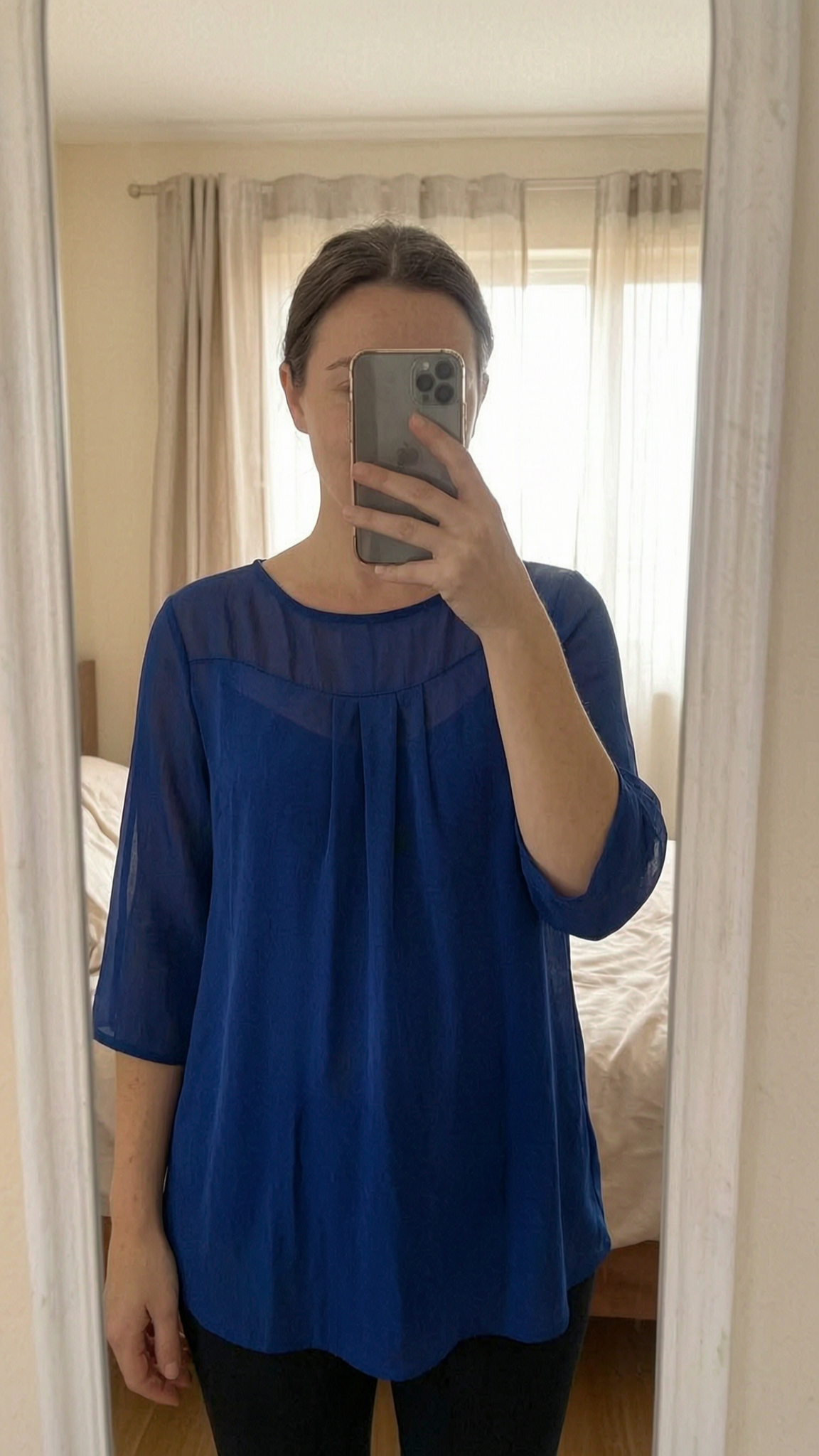 Blouse bleue manches 3/4 M - blusa donna in piedi, rivolto verso la fotocamera, in una camera da letto con luce soffusa e luce naturale che filtra dalla finestra.