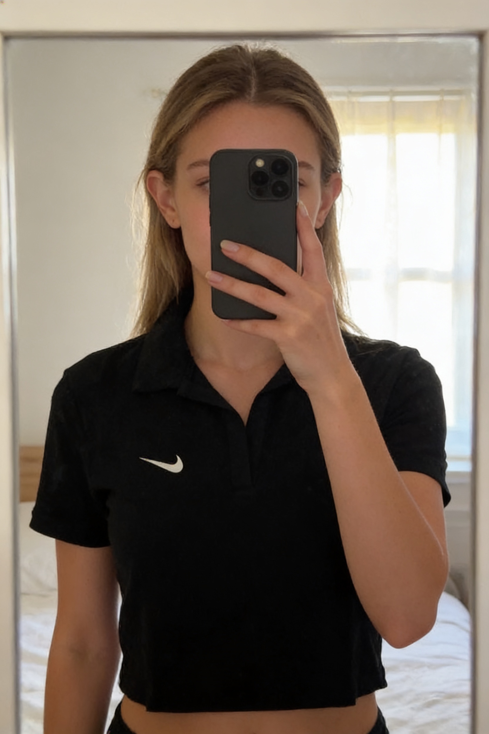 Polo court femme Nike L debout, face à la caméra, dans une chambre à la lumière douce, avec une lumière naturelle filtrant par la fenêtre.