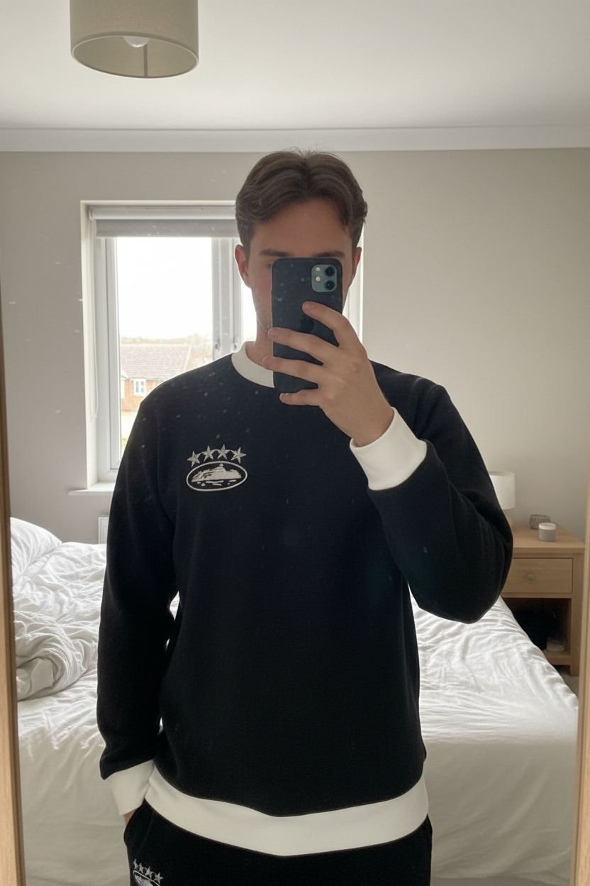 Pull Corteiz – Noir & Blanc debout, face à la caméra, dans une chambre à la lumière douce, avec une lumière naturelle filtrant par la fenêtre.