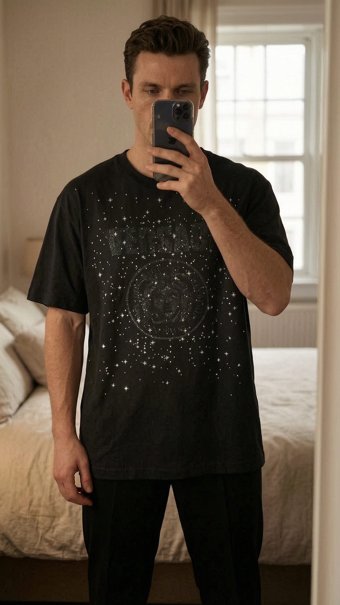 T-Shirt Versace Médusa debout, face à la caméra, dans une chambre à la lumière douce, avec une lumière naturelle filtrant par la fenêtre.