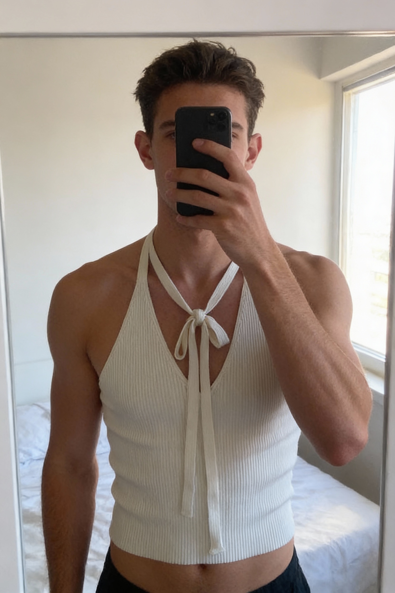 Top blanc côtelé dos-nu debout, face à la caméra, dans une chambre à la lumière douce, avec une lumière naturelle filtrant par la fenêtre.