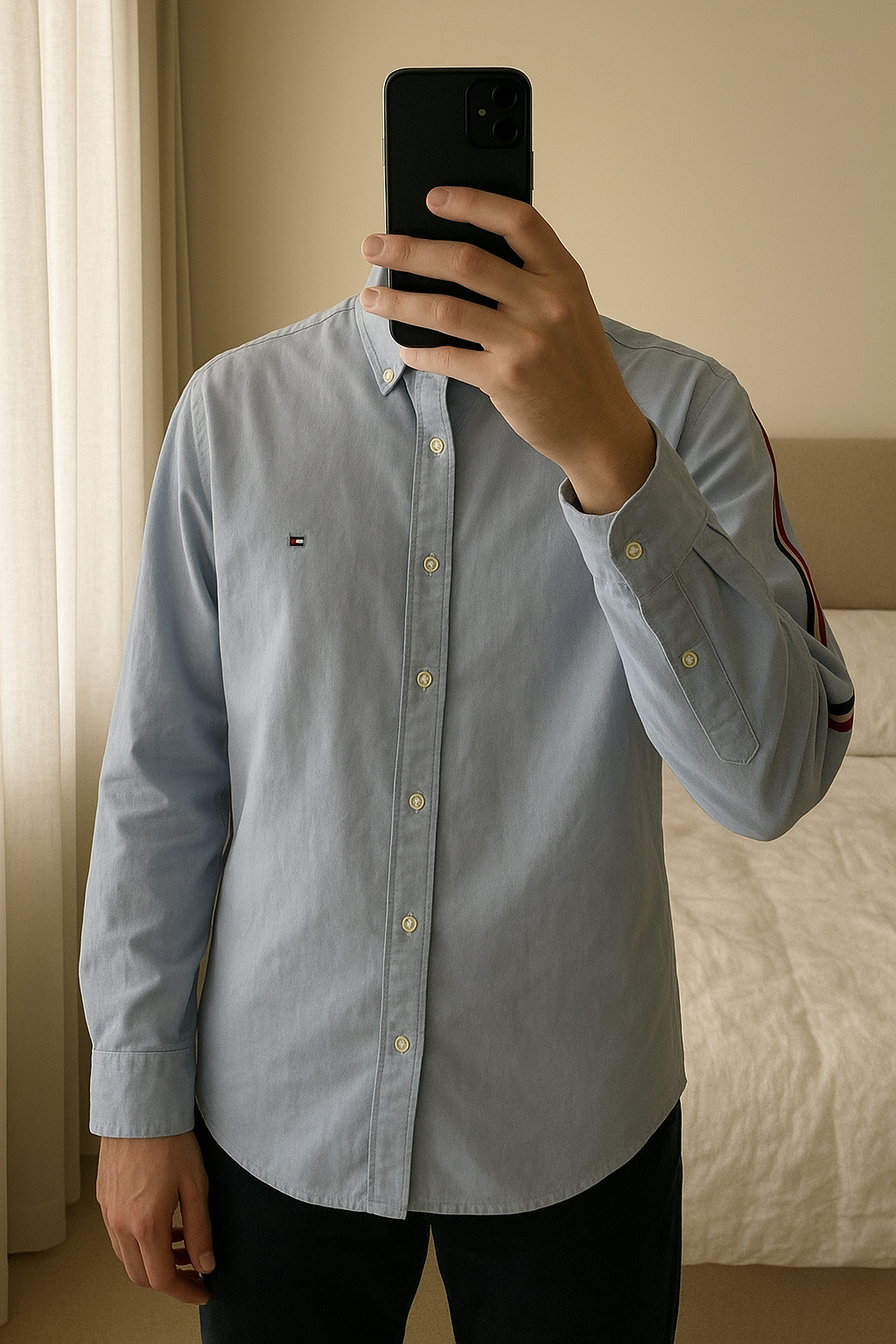 Chemise Tommy Hilfiger debout, face à la caméra, dans une chambre à la lumière douce, avec une lumière naturelle filtrant par la fenêtre.