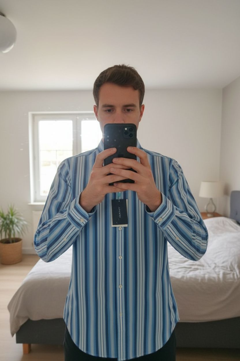 Chemise homme Gant debout, face à la caméra, dans une chambre à la lumière douce, avec une lumière naturelle filtrant par la fenêtre.