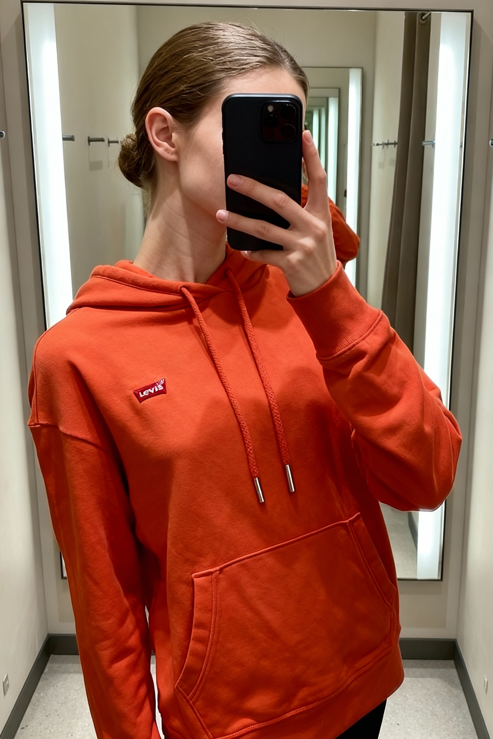 Una persona con Sudadera Levi’s naranja con capucha – talla S arqueando ligeramente la espalda para una pose dinámica, con tipo de cuerpo delgado, en un probador con estilo boutique, con espejo y decoración neutra.