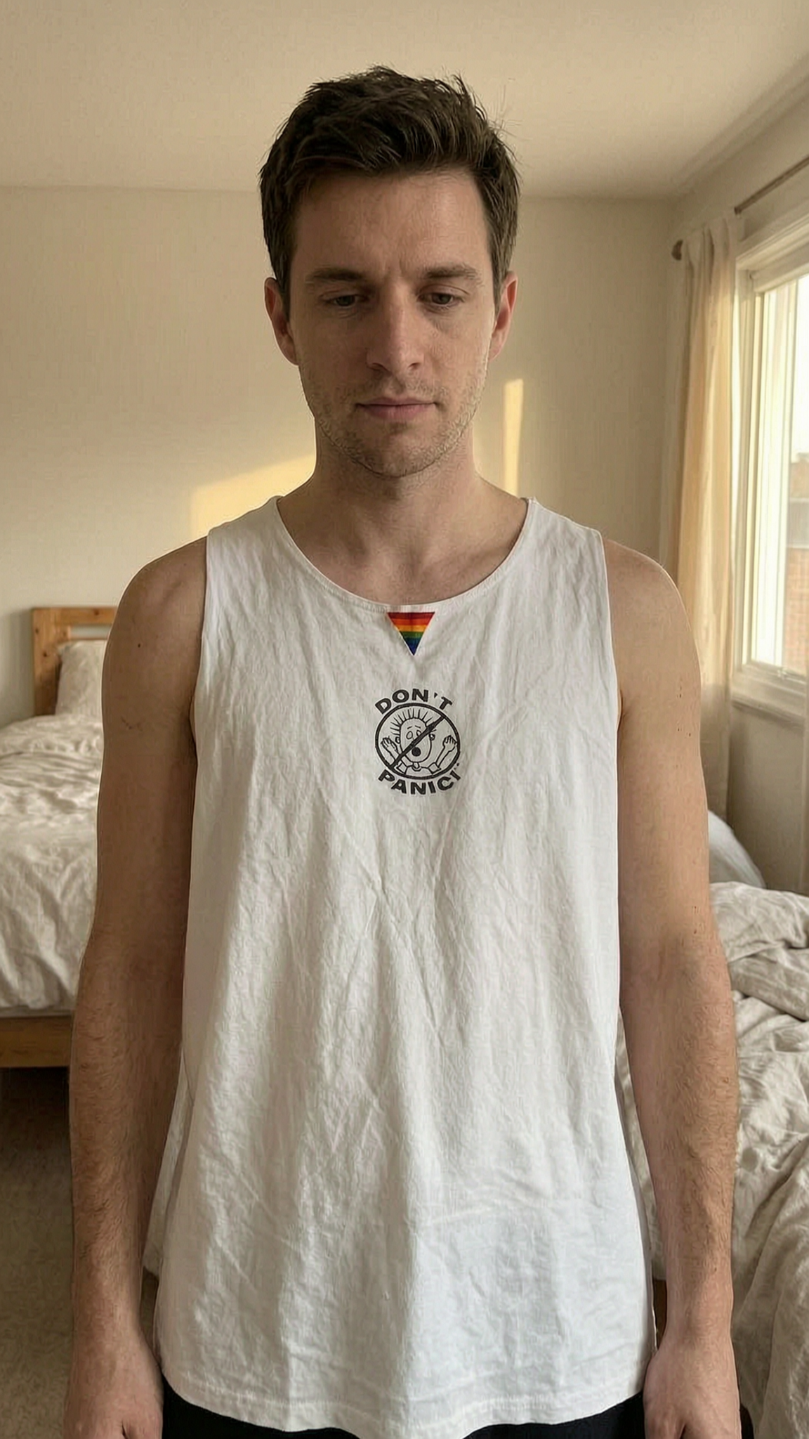 Men's Don't Panic white vest with Pride triangle. de pie, mirando a la cámara, en un dormitorio con luz suave y natural filtrándose por la ventana.