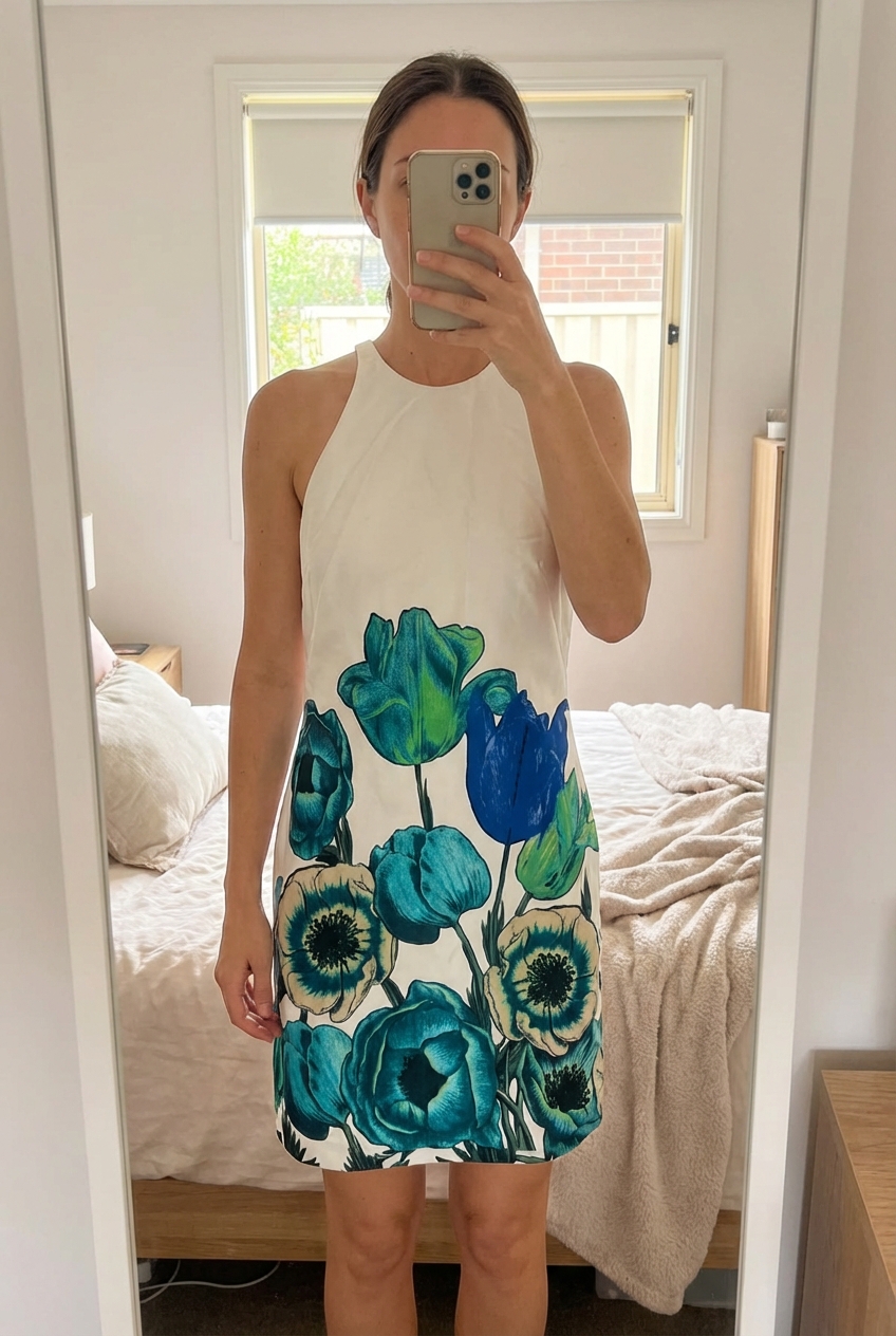 Une personne en Robe blanche à fleurs bleues et vertes – Taille XS, en bon état debout, face à la caméra, avec une silhouette fine, dans une chambre à la lumière douce, avec une lumière naturelle filtrant par la fenêtre.